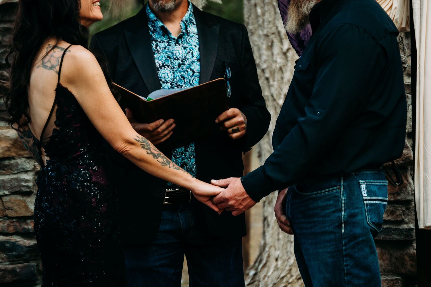 Couple holds hands during intimate outdoor wedding ceremony with officiant holding book.