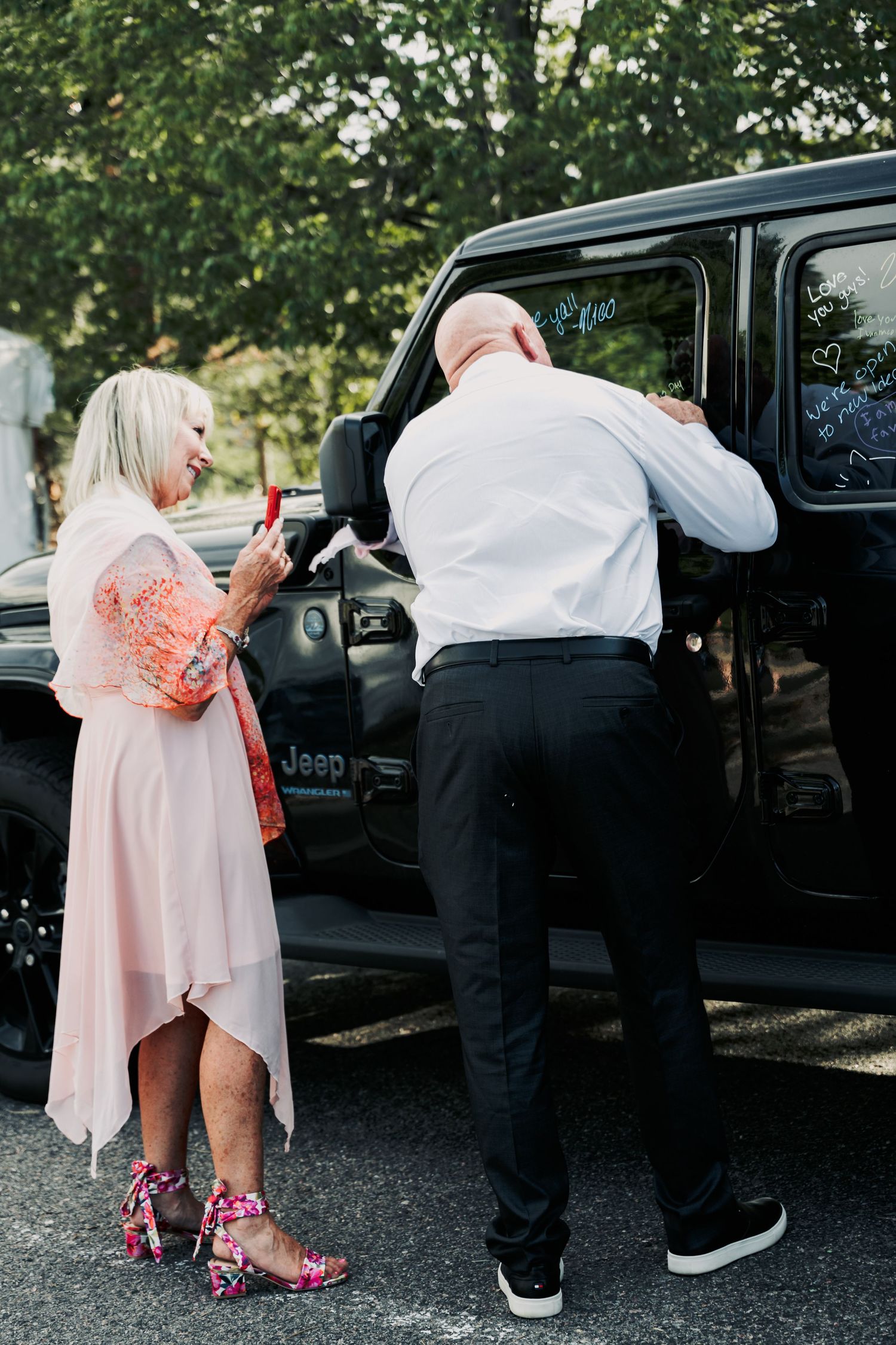 Couple writing on black Jeep Wrangler windows at wedding reception outdoor parking lot celebration.