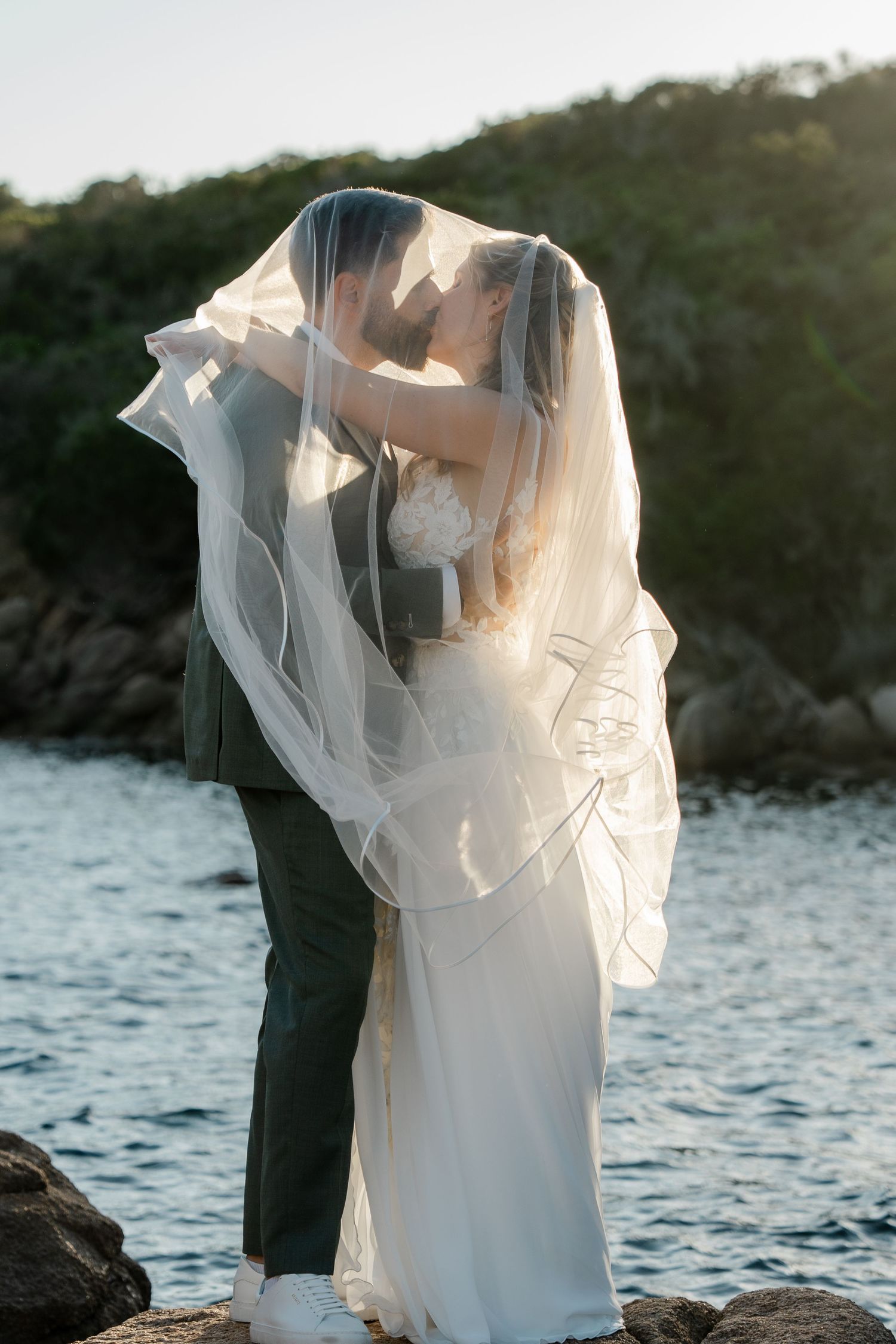 Bride and groom kissing under flowing veil on rocky shore with ocean and green hills at sunset.