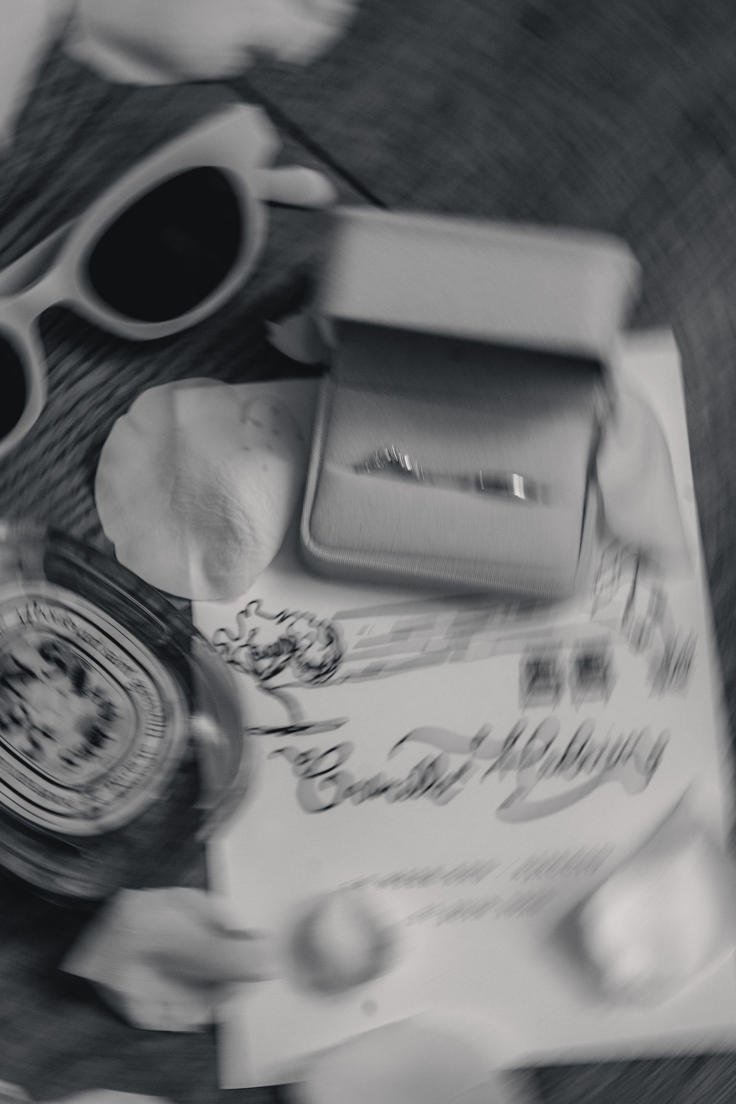 Black and white flat lay of an engagement ring in a box, sunglasses, candle, and magazine on a table.