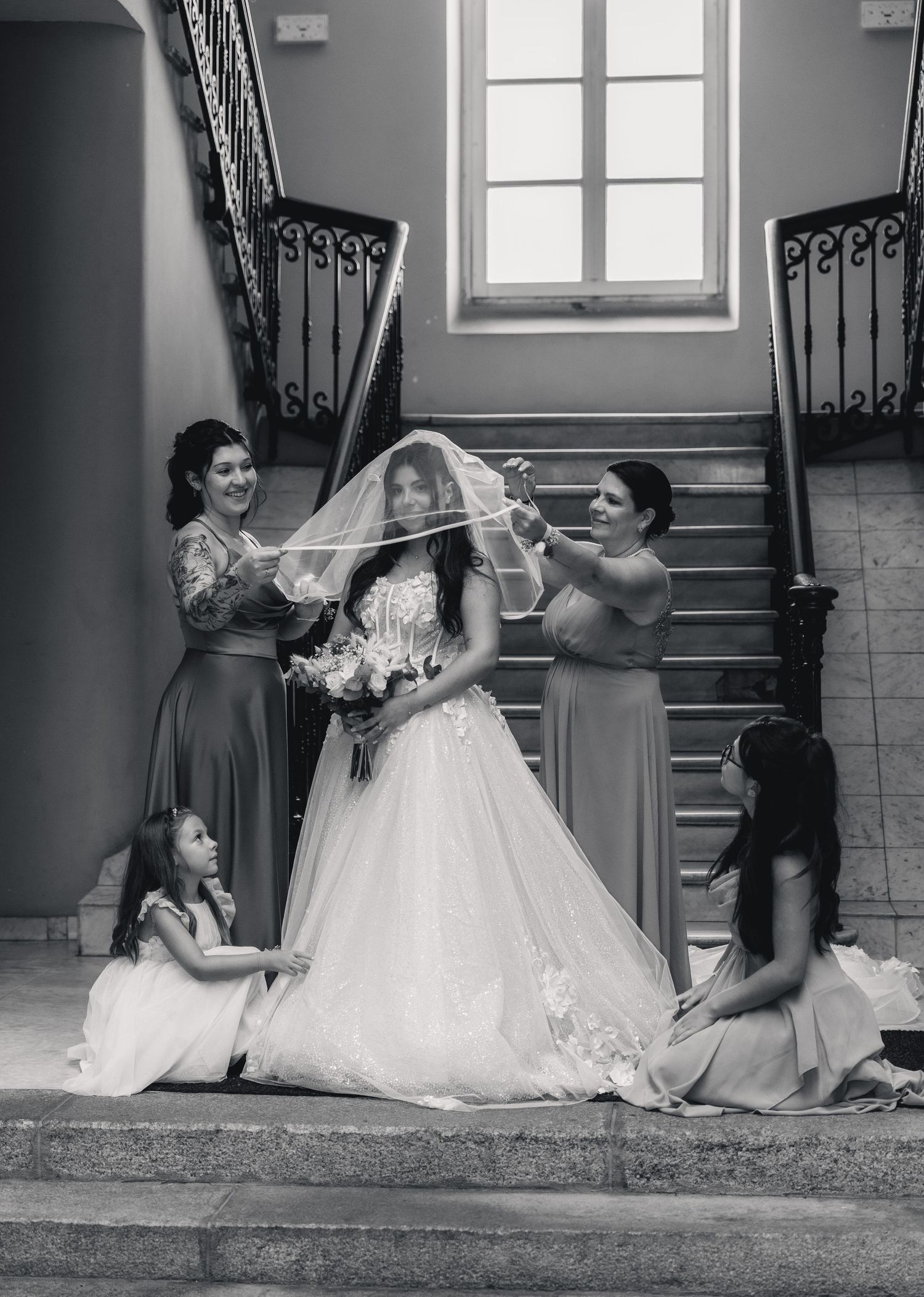 Black and white photo of a bride in a white gown and veil surrounded by bridesmaids on an elegant staircase.