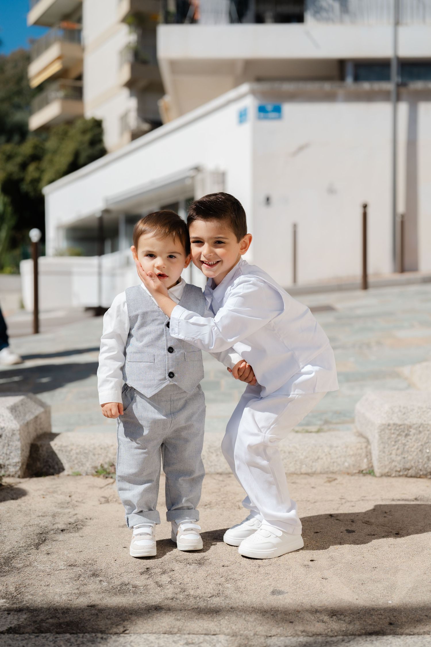 Two young children in matching white outfits play and laugh together on a sunny outdoor patio.
