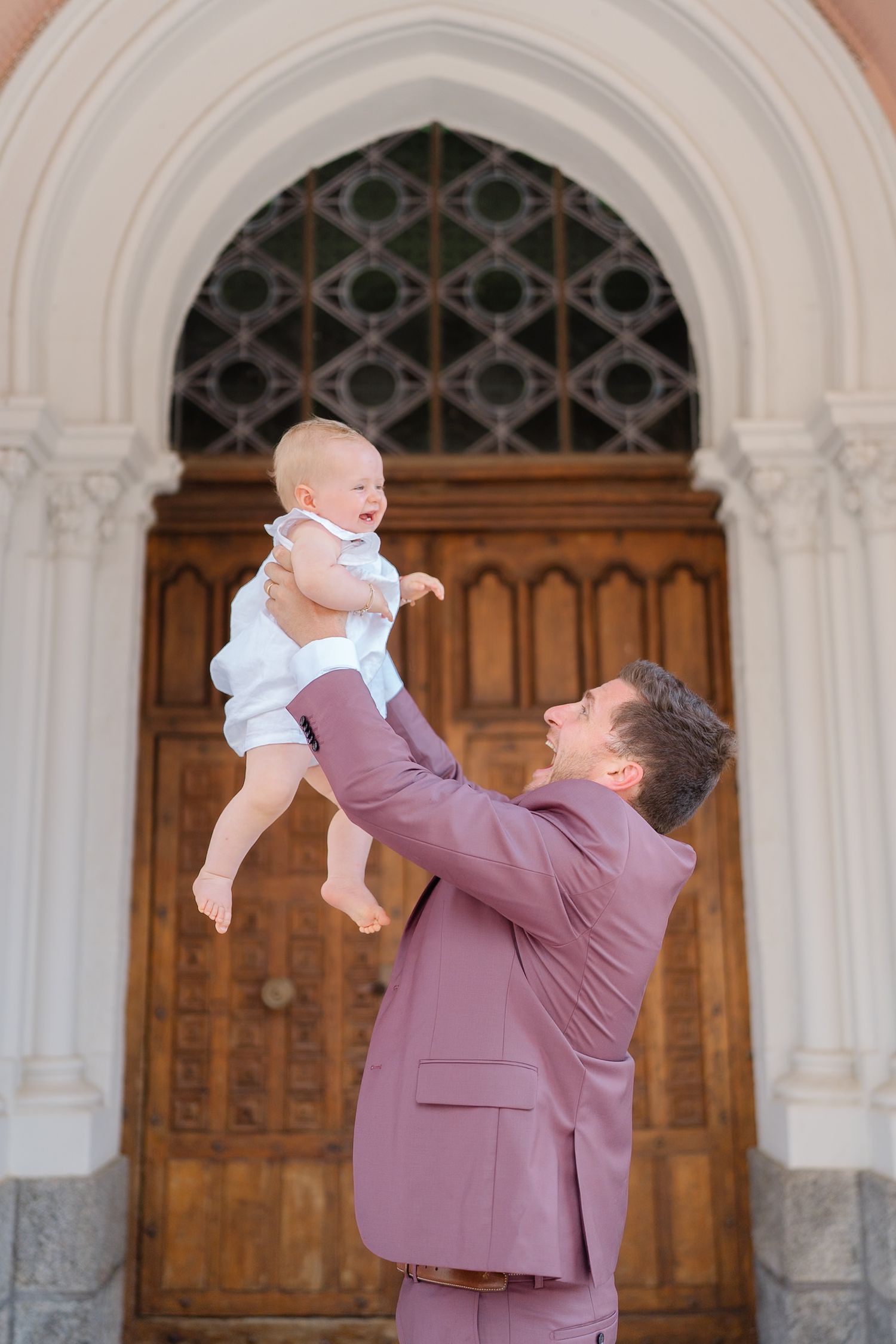 A parent in a mauve suit lifts and plays with a baby wearing white in front of ornate wooden church doors.