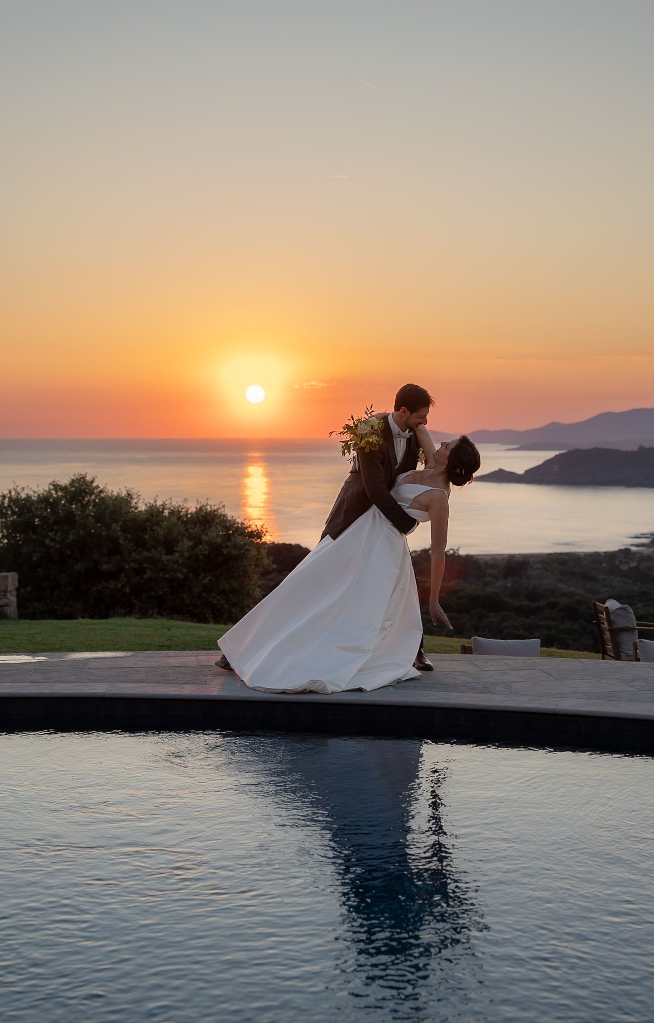Bride and groom share a romantic dip kiss by an infinity pool at sunset with ocean views.