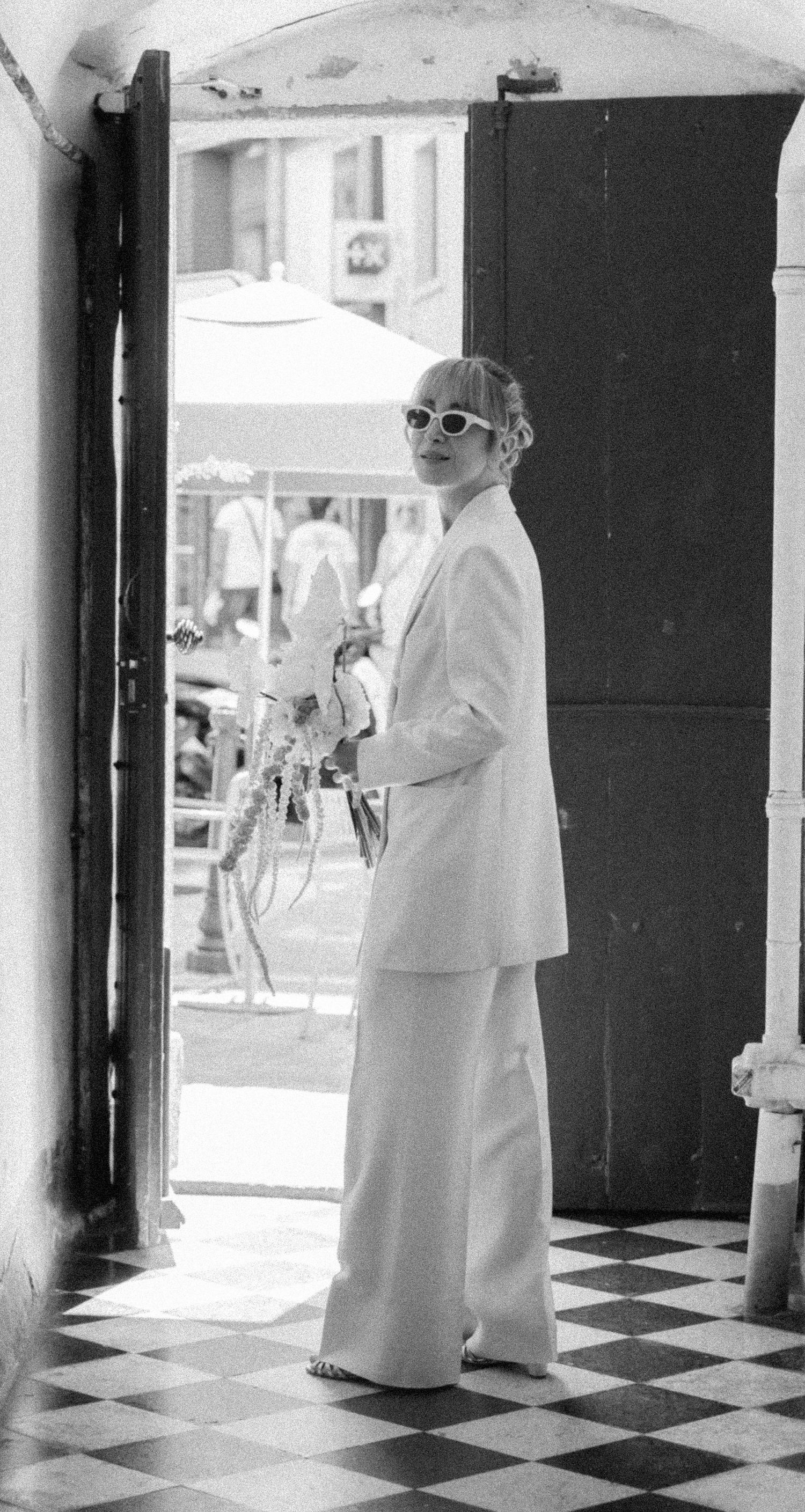 Bride in white pantsuit and sunglasses holding bouquet stands in doorway on checkered floor, black and white photo.