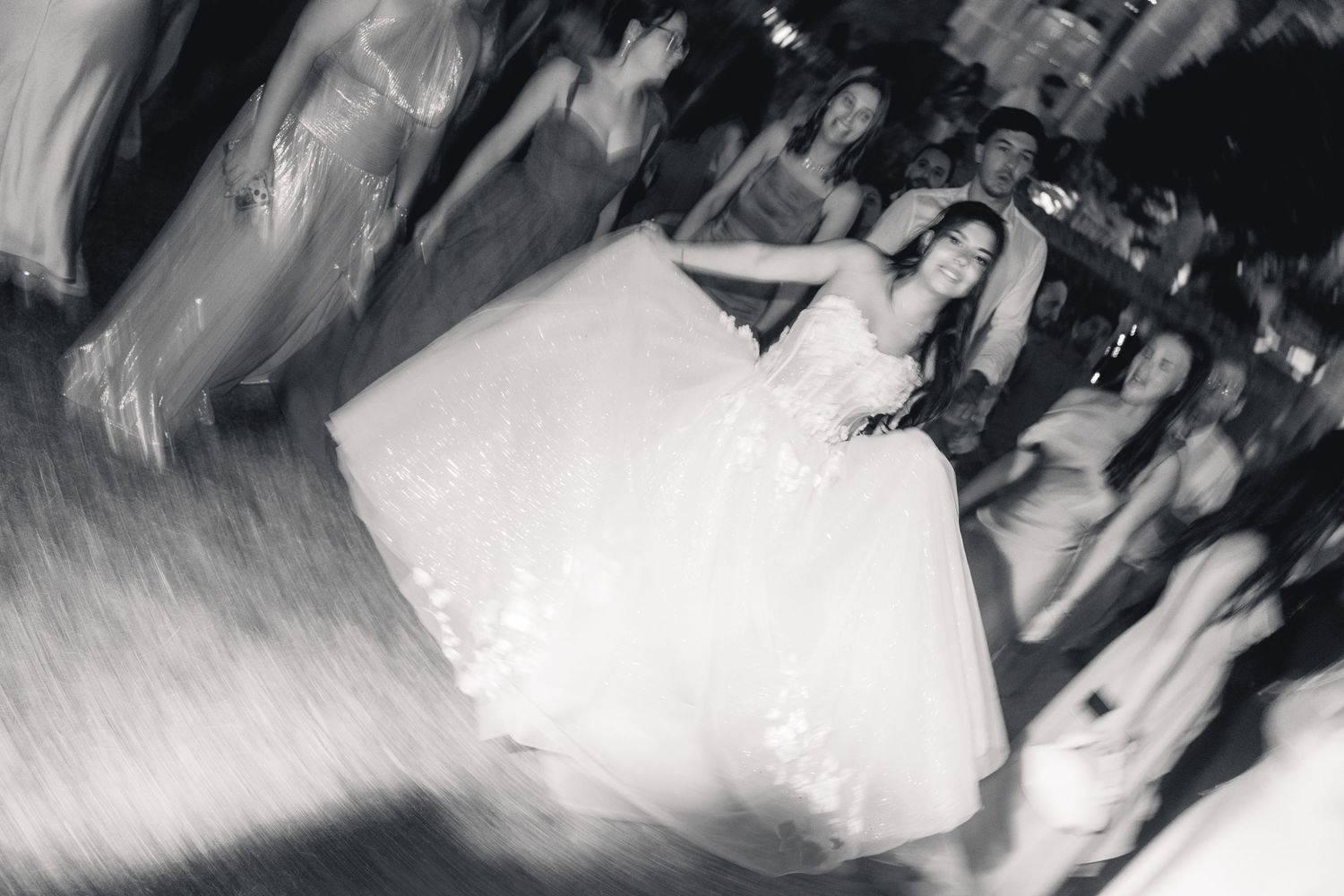 Black and white photo of a bride dancing joyfully in her lace wedding gown surrounded by guests at a reception.