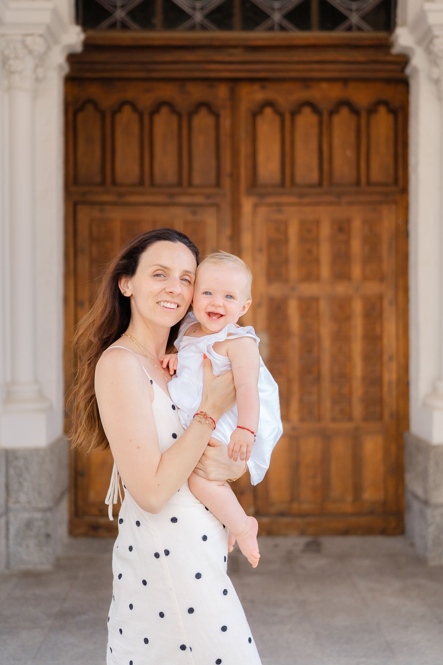 A polka dot dress-wearing woman shares happy moments with a baby in front of decorative wooden doors.