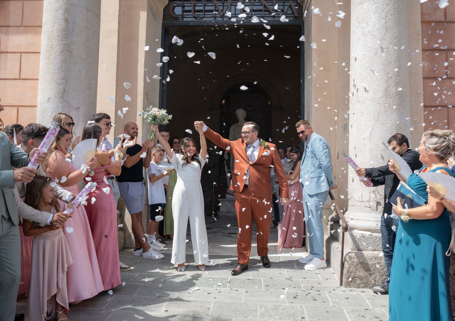 Joyful newlyweds exit church amid confetti shower, bride in white jumpsuit, groom in rust suit, guests cheering.