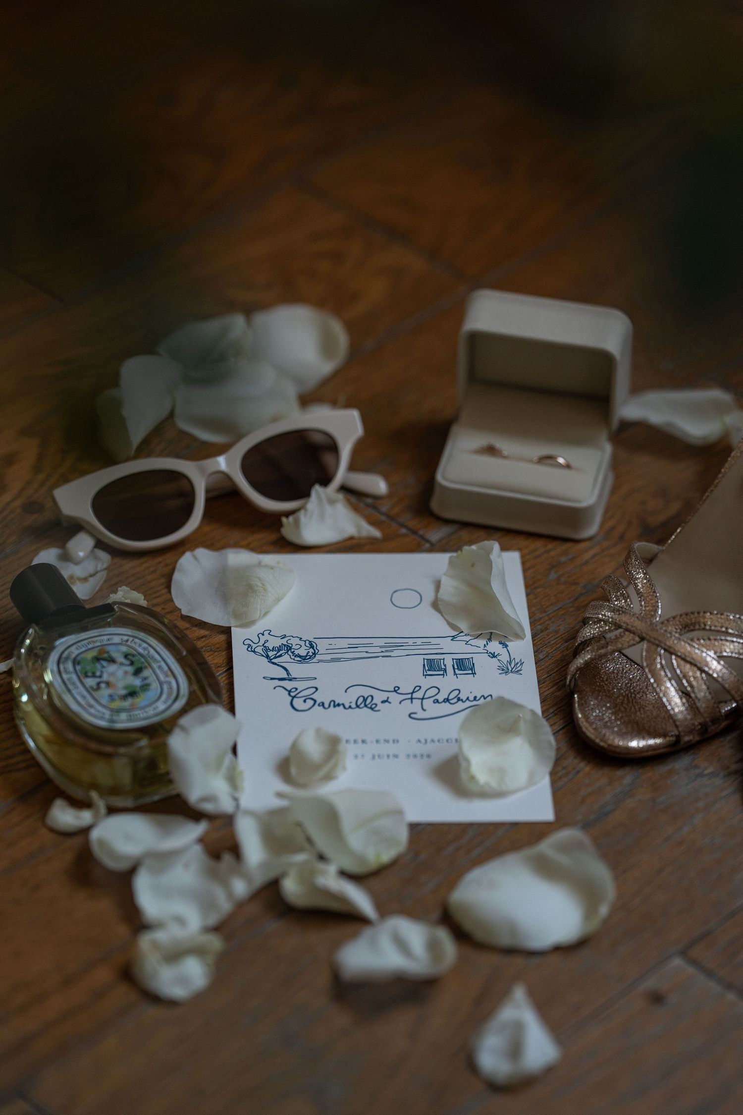 Wedding flat lay with ring box, sunglasses, rose petals, perfume, invitation, and gold heels on wooden surface.