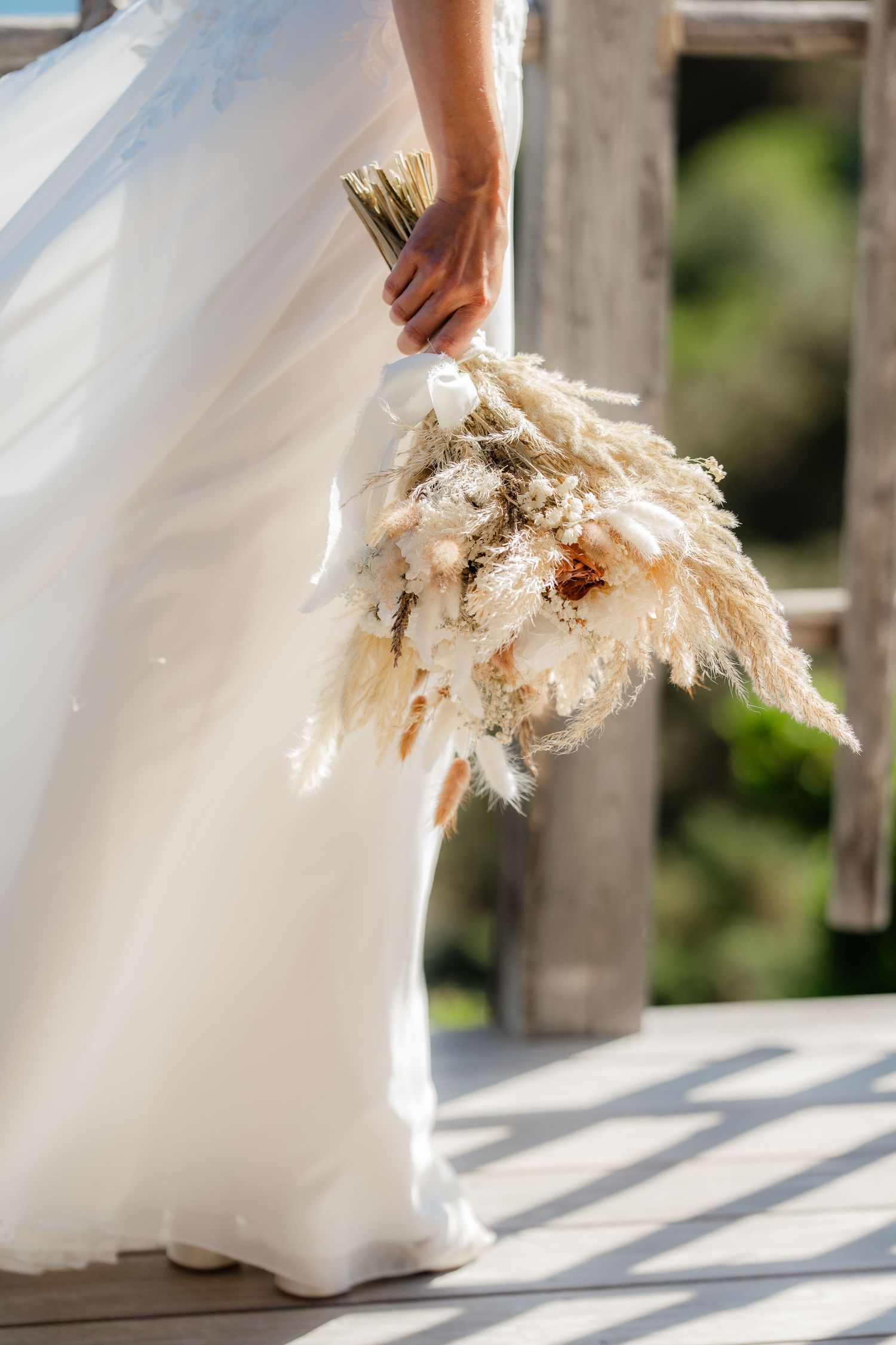 Bride in white gown holding a boho dried pampas grass wedding bouquet outdoors in natural sunlight.
