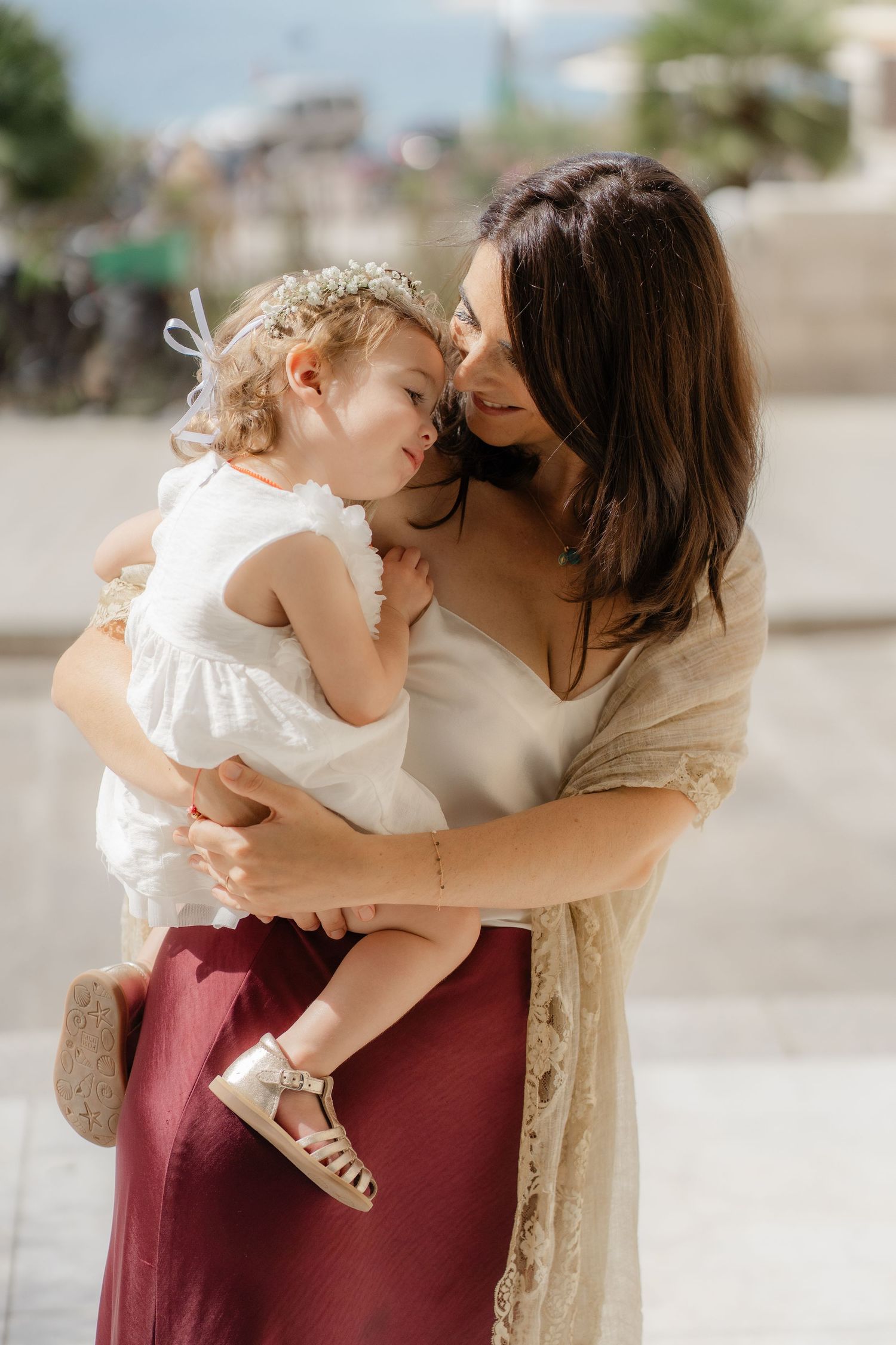 A tender moment between parent and child as they embrace outdoors, the child wearing a white dress and the parent in burgundy.