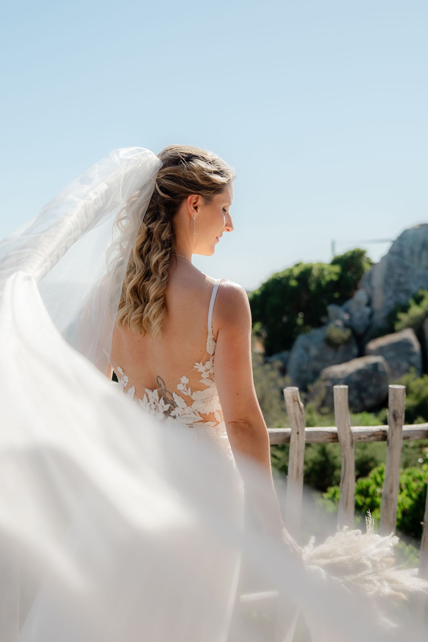 Bride in a lace backless wedding dress with flowing veil blowing in the wind, standing outdoors on a sunny day.