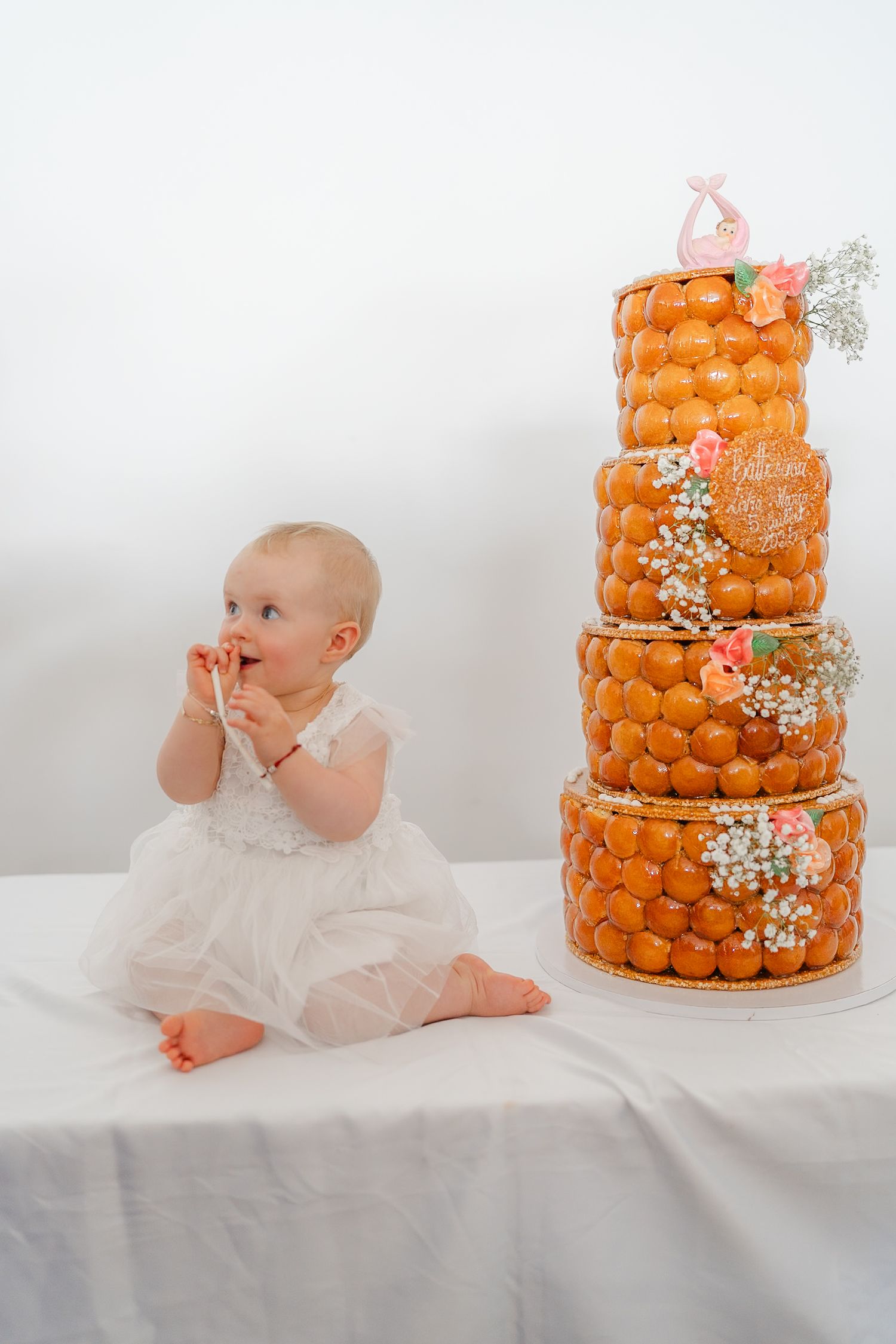 Baby in white dress sitting next to a three-tiered orange croquembouche cake decorated with baby's breath flowers.