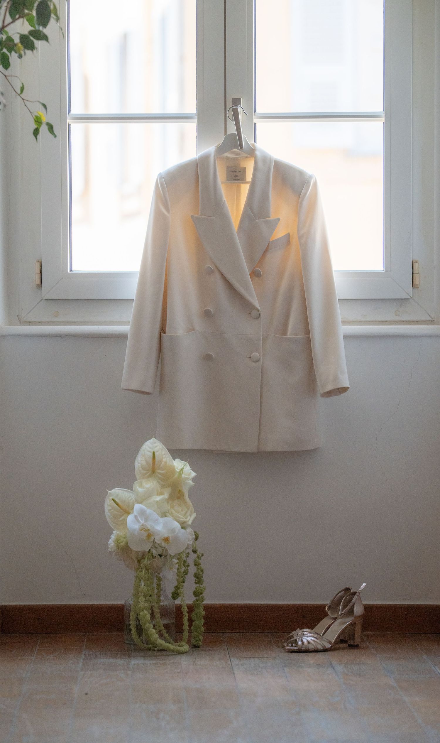 White double-breasted blazer hanging by window with white floral bouquet and silver heels on floor below.