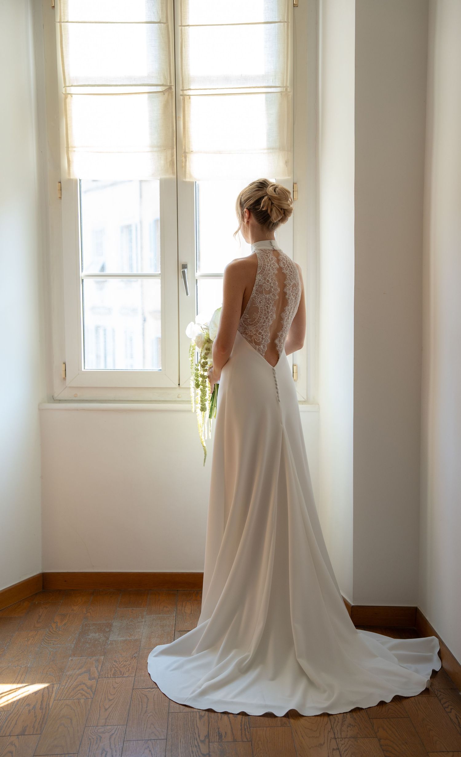 Bride in elegant backless lace halter wedding gown with train holds bouquet while gazing out sunlit window.