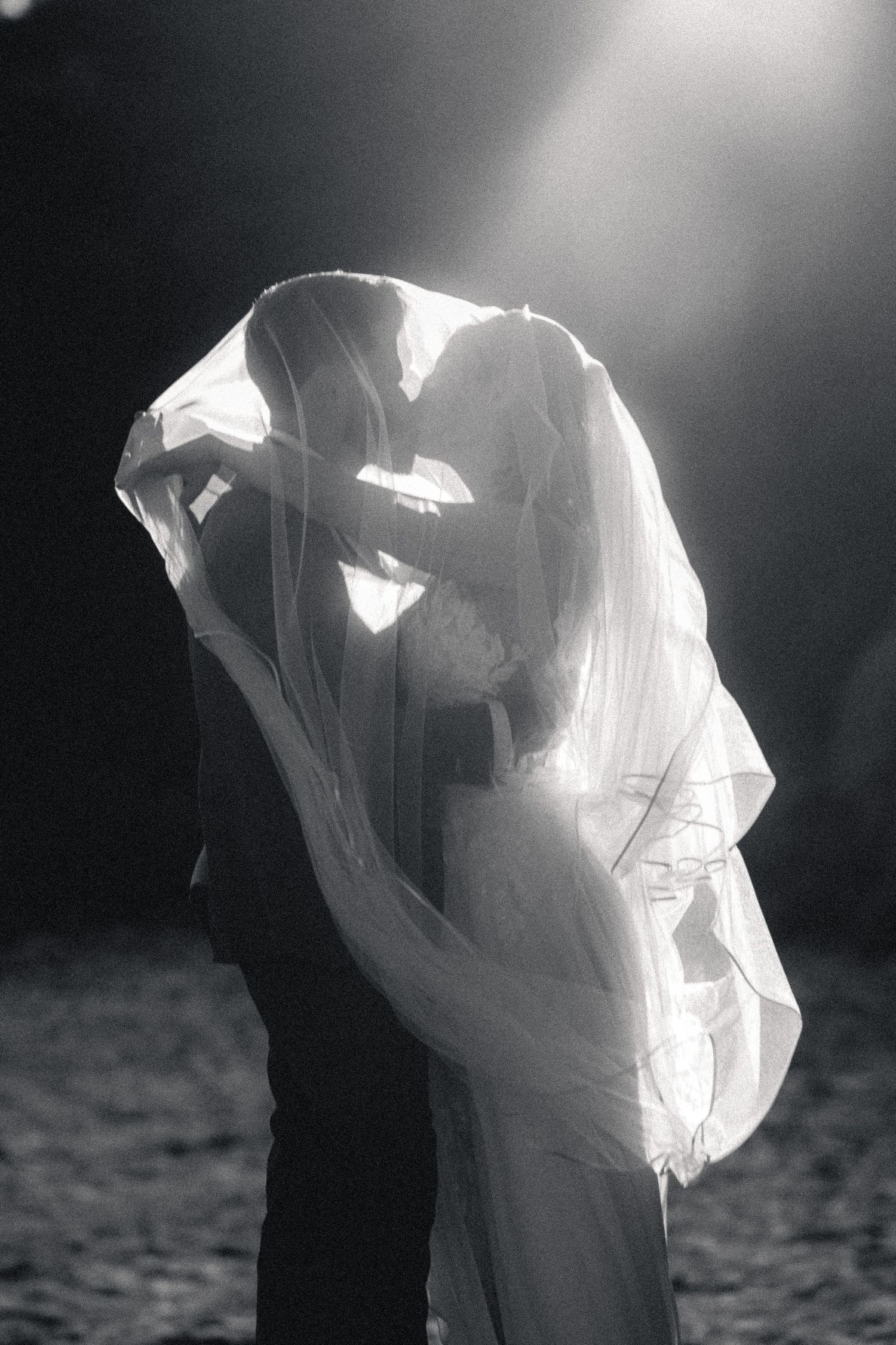 Black and white photo of bride and groom kissing under flowing veil with dramatic backlight.