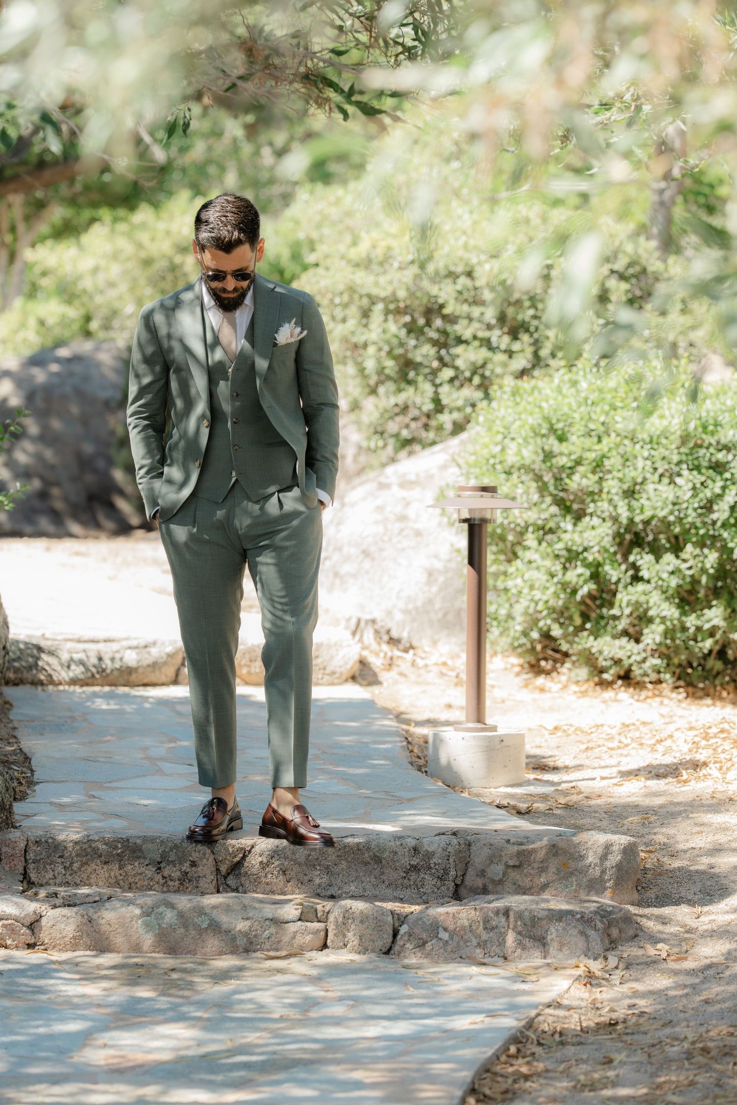 Man in sage green three-piece suit with sunglasses standing on stone steps in a sunlit garden.