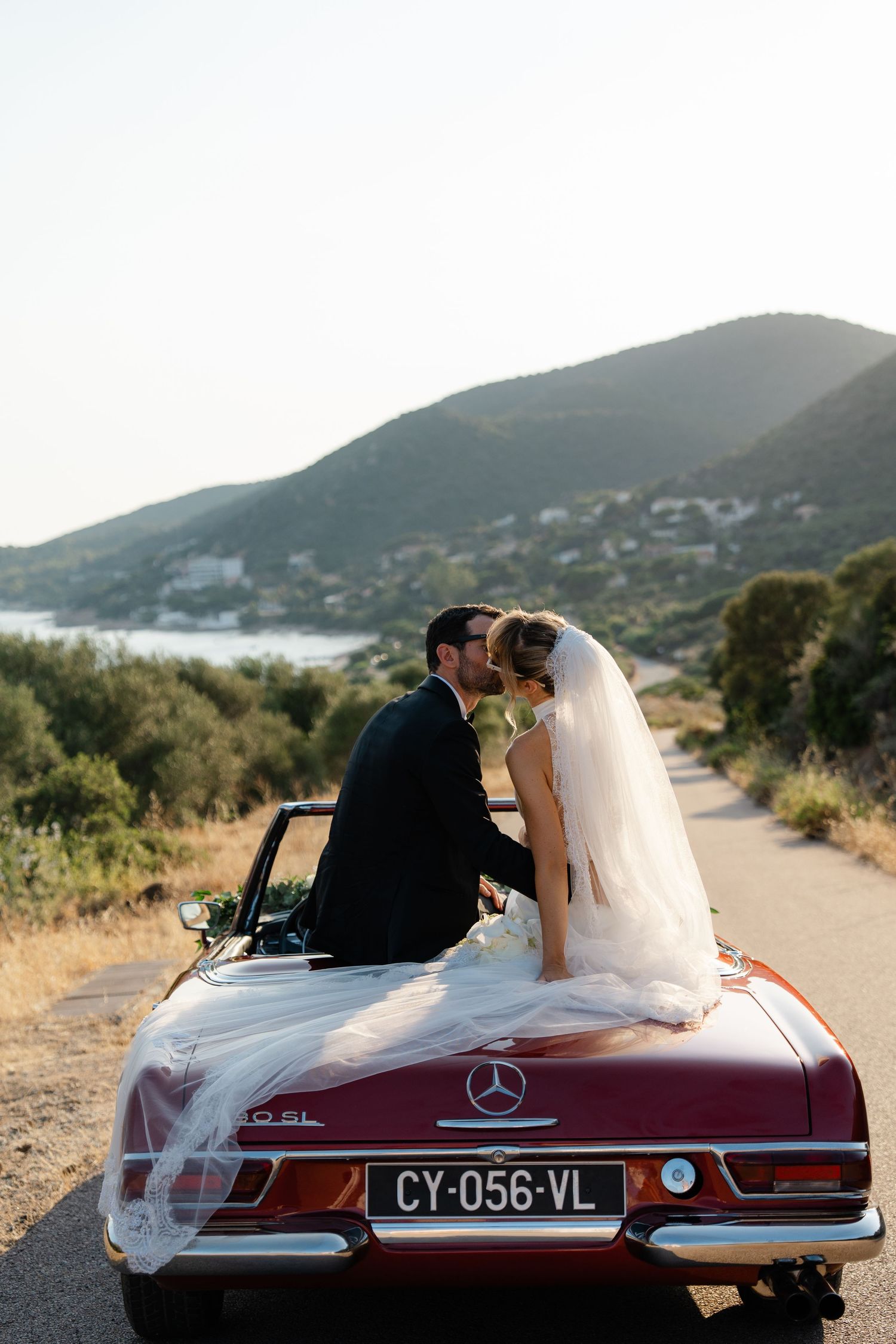 Bride and groom kissing in a vintage red Mercedes convertible on a scenic road with green hills and water in the background.
