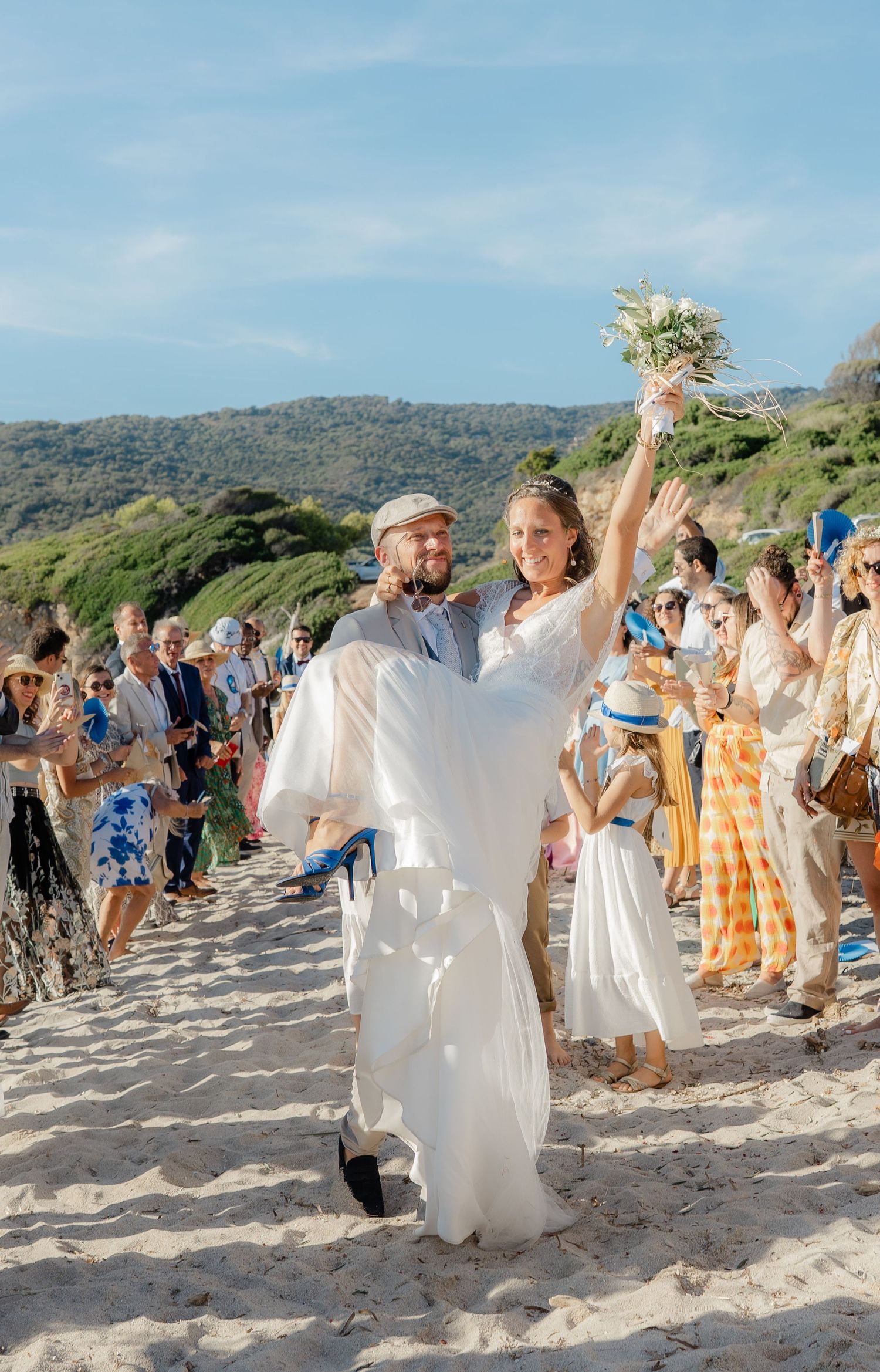 Wedding celebration on a sunny beach with guests cheering as couple celebrates.