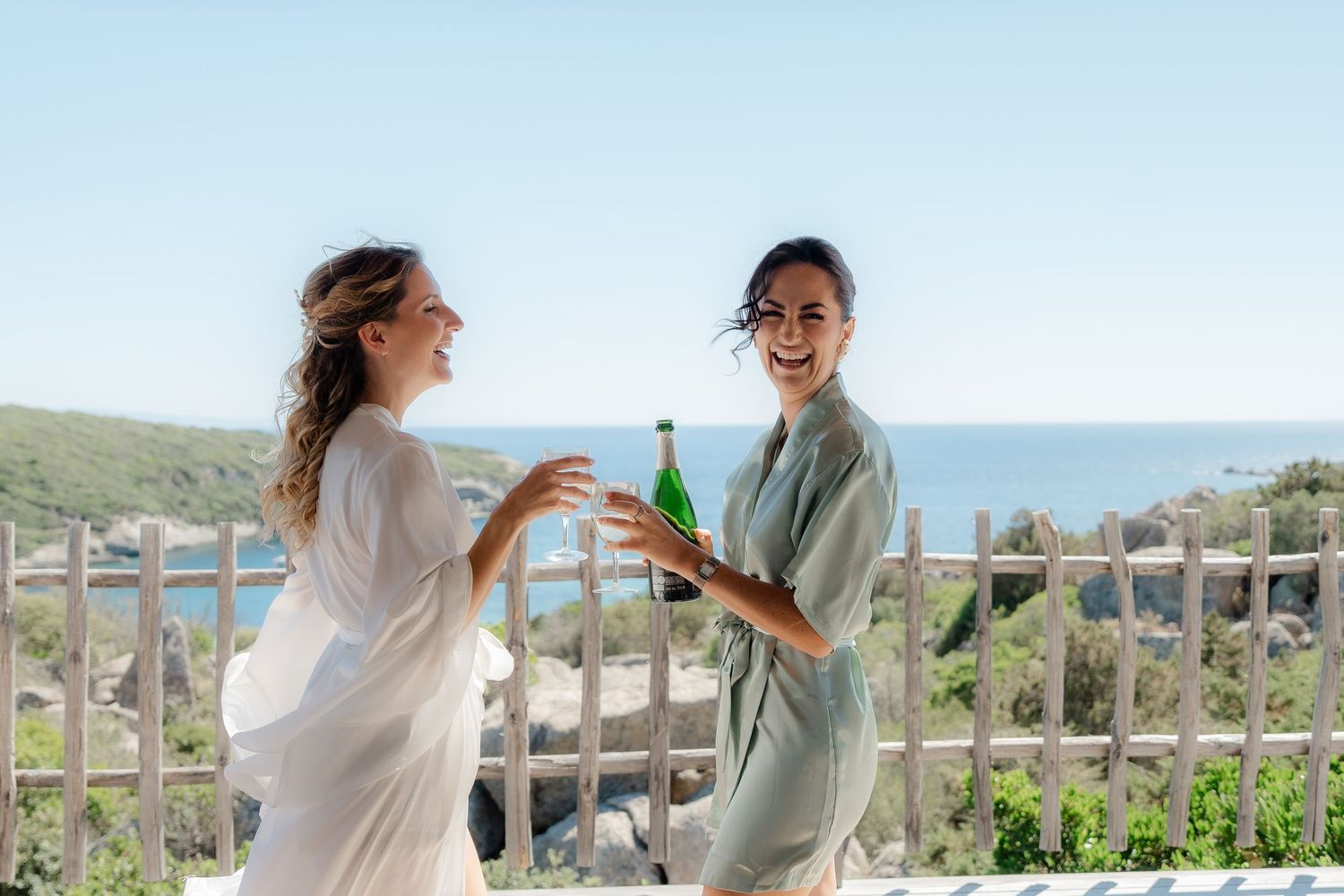 Two women celebrating outdoors with champagne, smiling by a scenic ocean view on a sunny day.