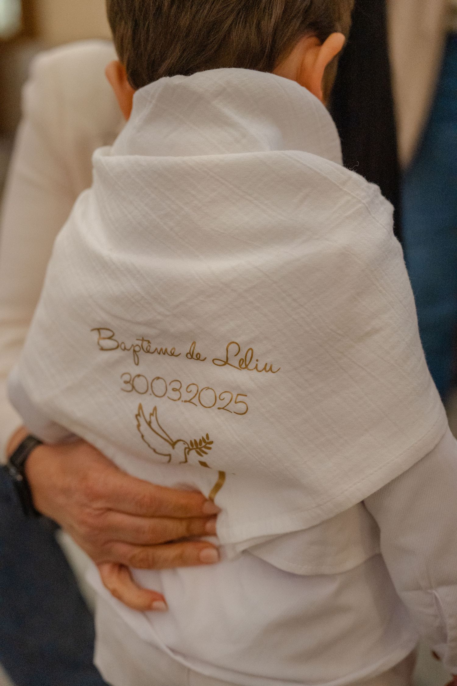 Close-up of white baptismal garment with embroidered date 30.03.2025 and religious symbol being worn during ceremony.