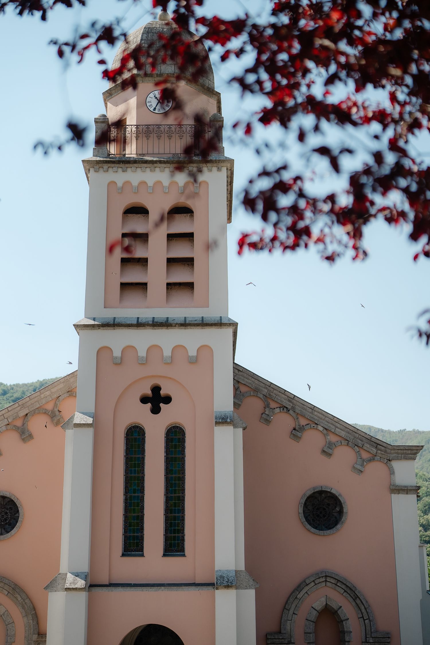 Pink church facade with tall bell tower and green stained glass windows framed by red autumn leaves.