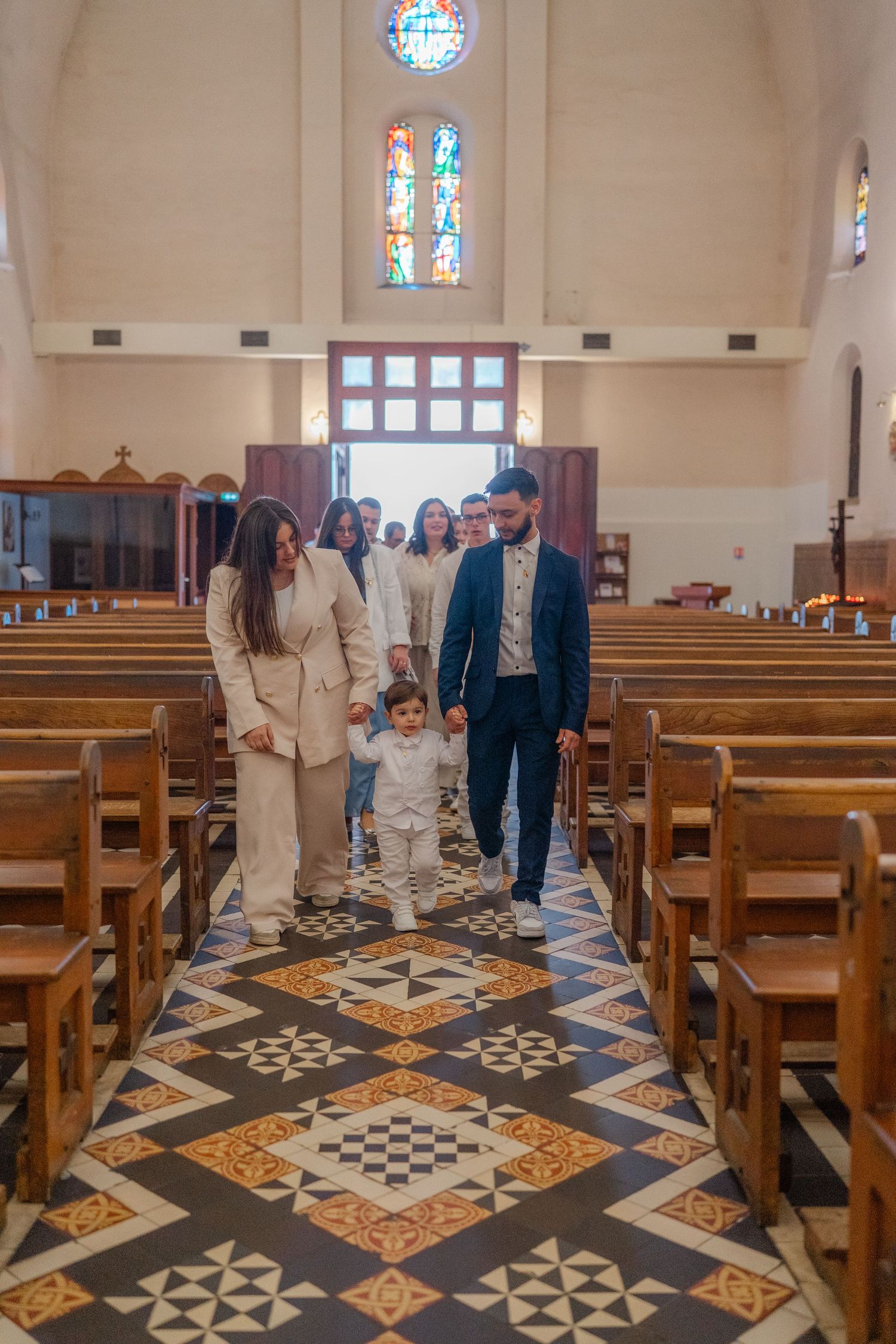 Church interior with ornate tiled floor pattern and wooden pews arranged in rows beneath stained glass windows.