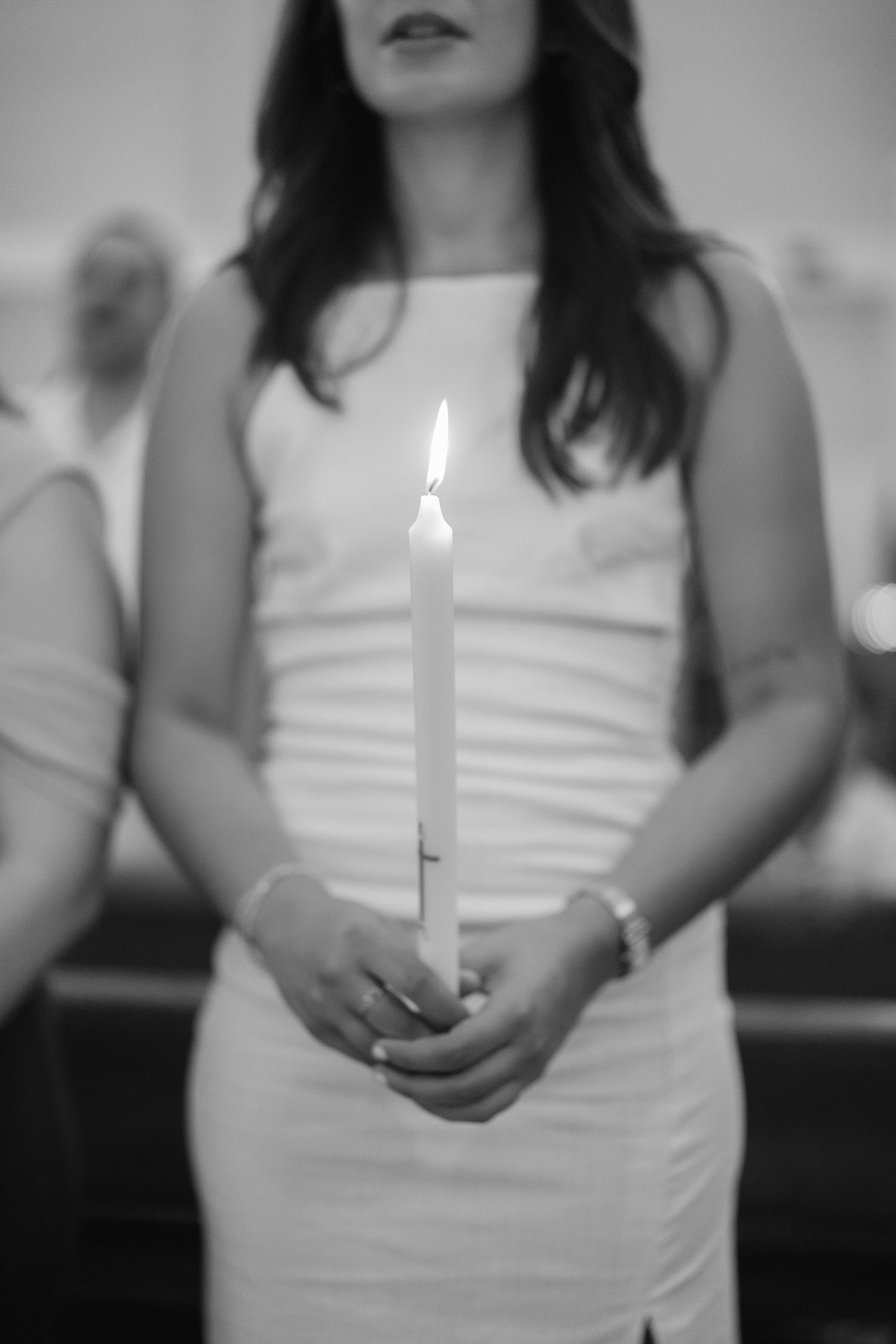 A person in a white sleeveless dress holds a lit candle in a dimly lit setting.