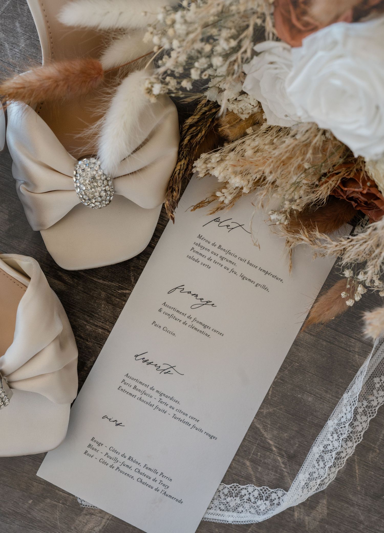 Elegant wedding flat shoes with bow and rhinestone detail beside a menu card and dried floral bouquet.