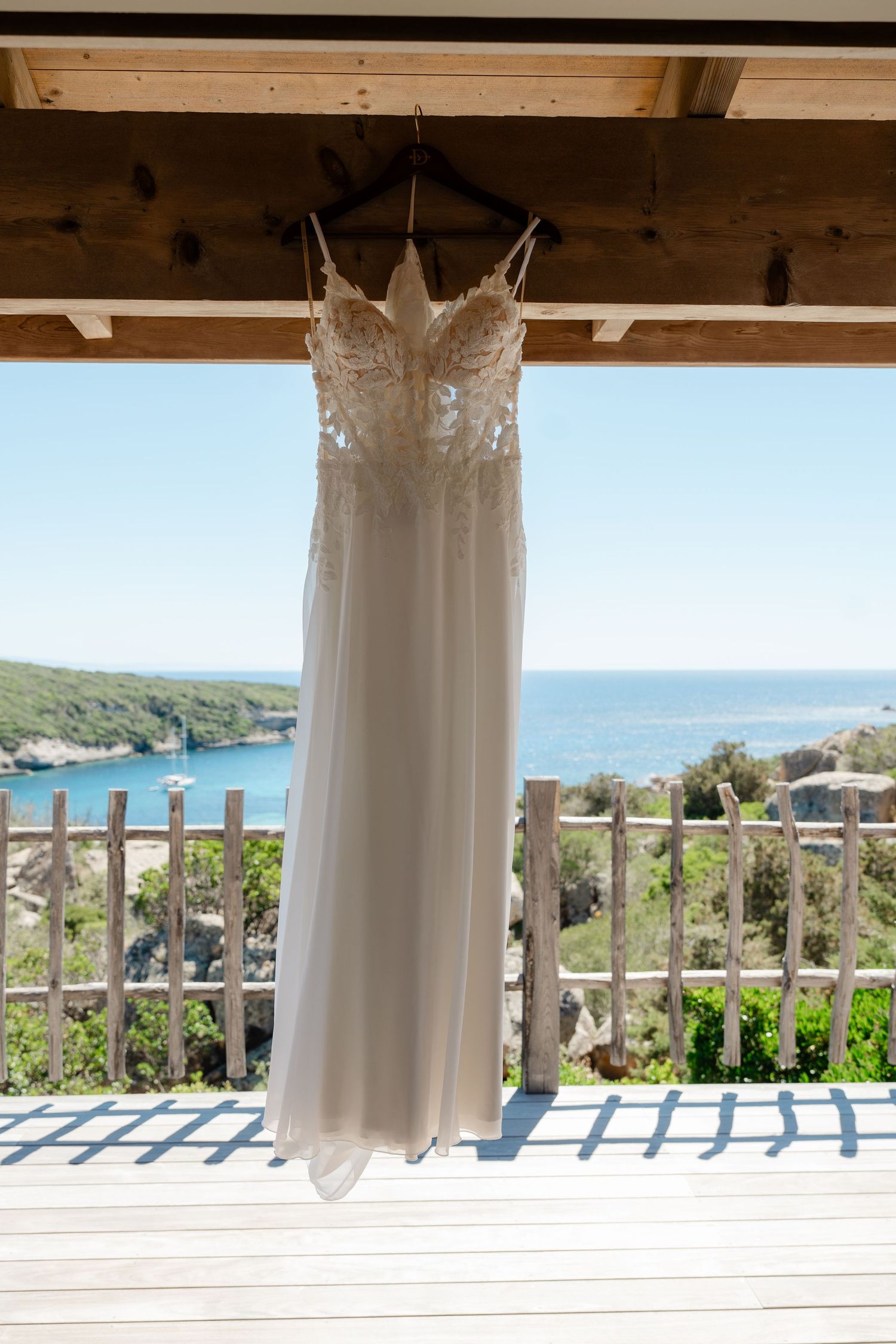 Elegant white lace wedding dress hanging on a wooden beam with a scenic ocean and greenery backdrop.