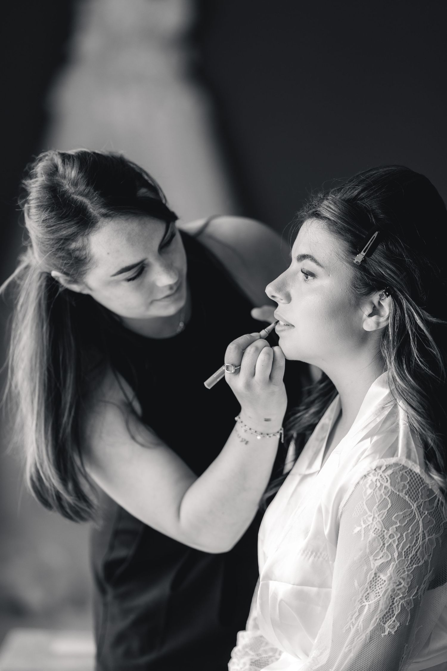 Black and white photo of a makeup artist applying lipstick to a bride in a white lace robe before her wedding.