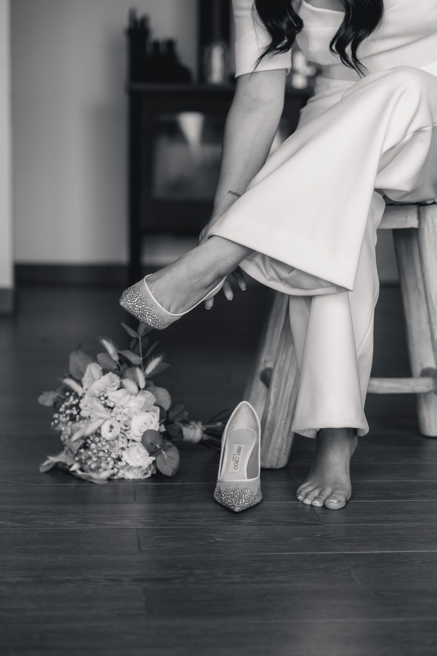 Black and white photo of a bride in white dress putting on sparkly heels beside a floral bouquet on wooden floor.