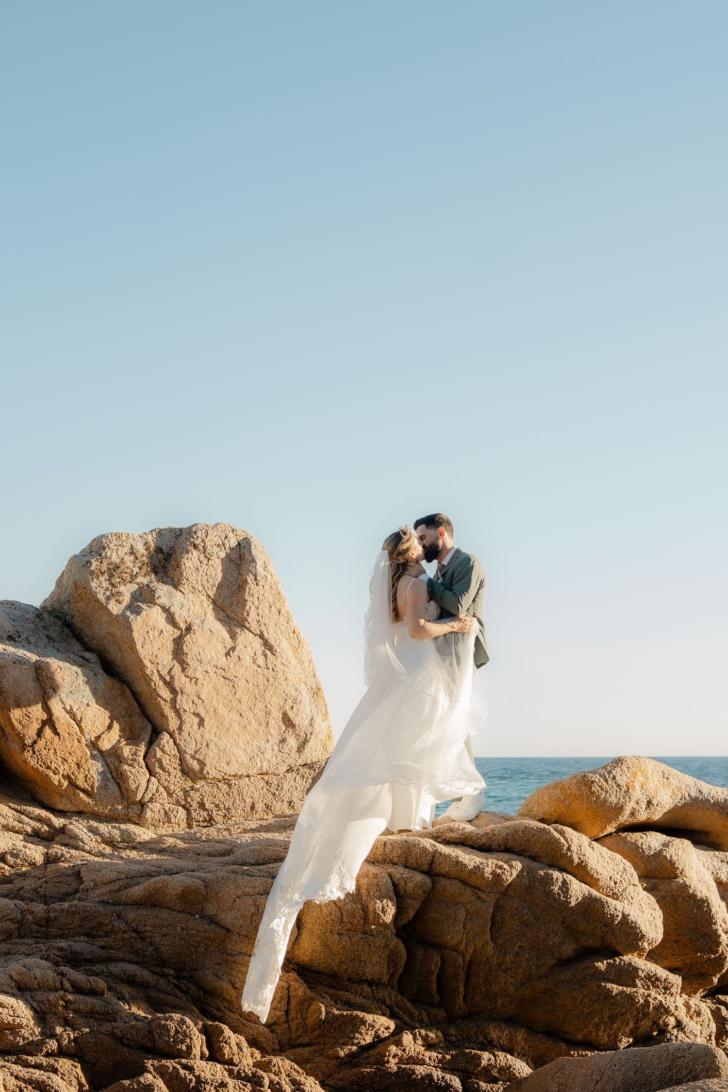 A bride and groom share a romantic kiss on rocky coastal cliffs with the blue ocean and clear sky behind them.