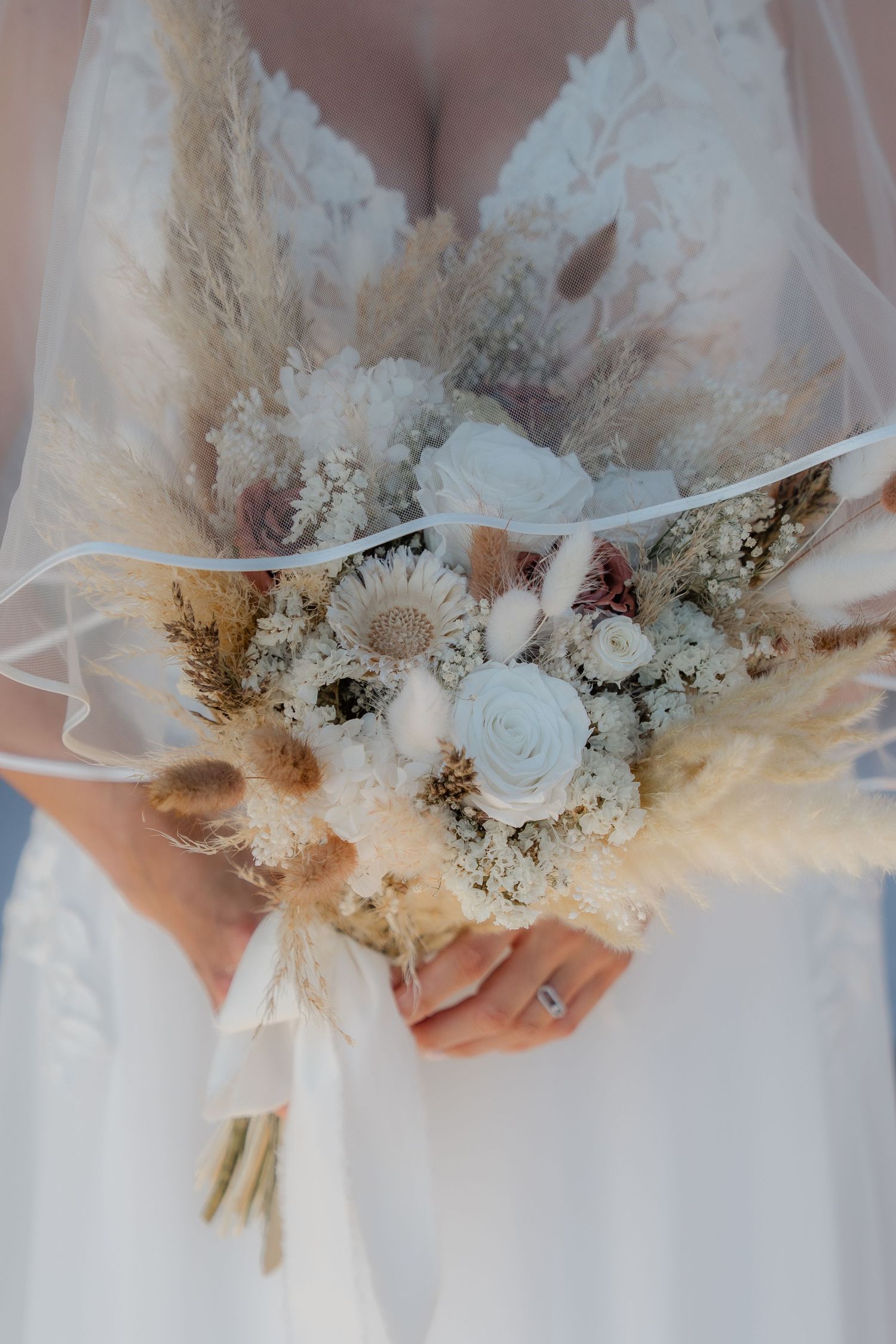 Bride holding a boho dried flower wedding bouquet with white roses, pampas grass, and wildflowers, wearing a lace gown.