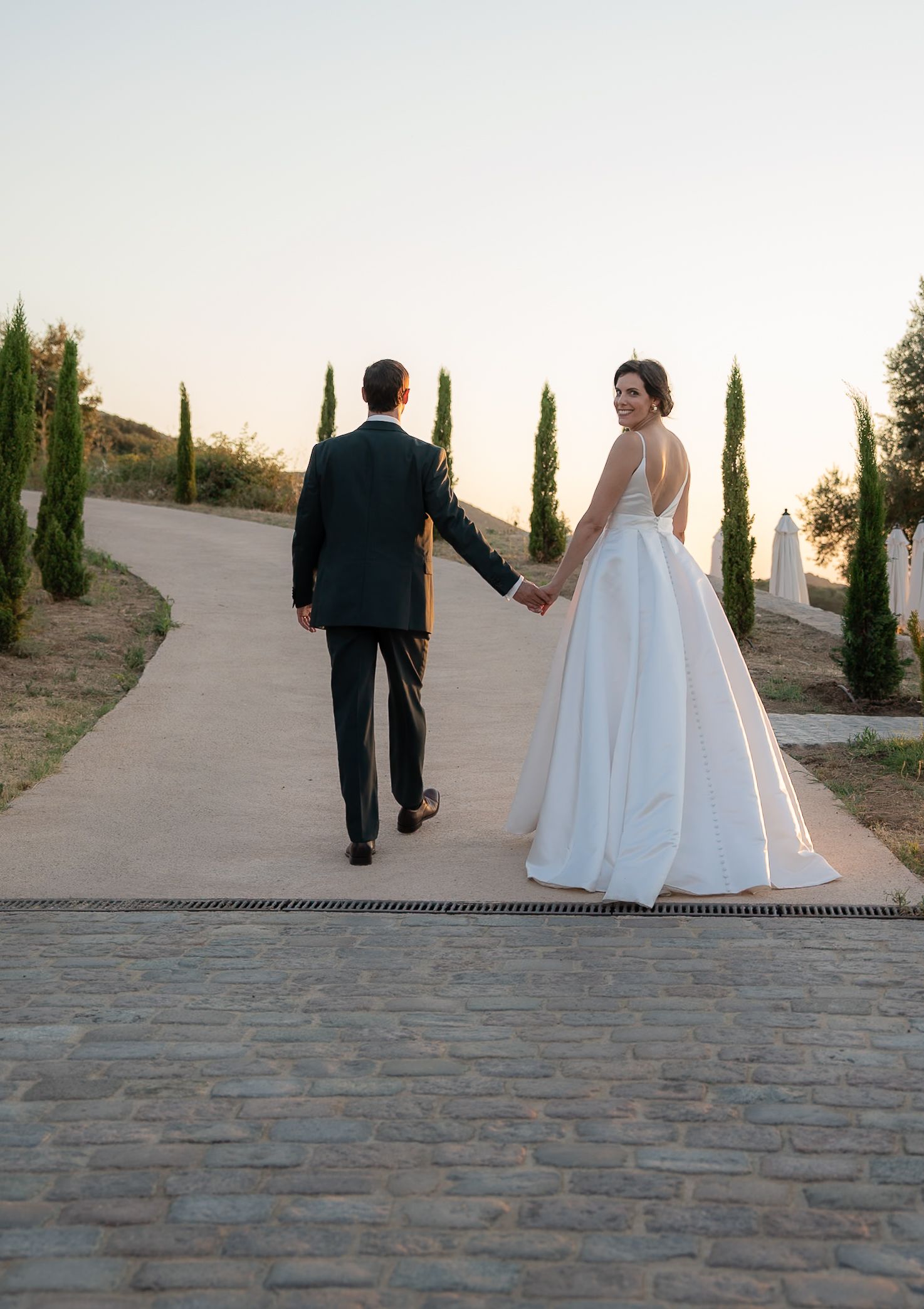 Bride in white gown and groom in dark suit walk hand-in-hand down a cobblestone path lined with cypress trees.