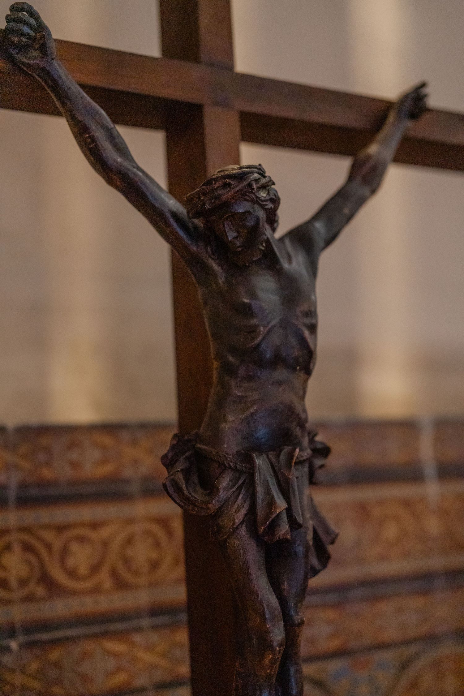 Close-up of a dark bronze crucifix showing Jesus on the cross with ornate wooden background details.