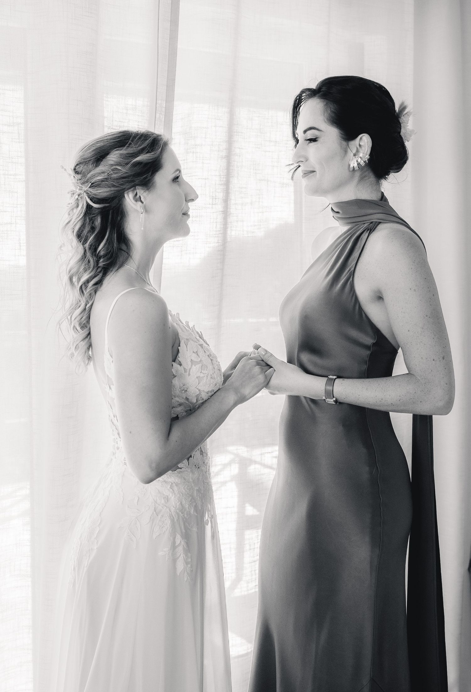 Black and white photo of a bride in a lace gown holding hands with a bridesmaid in an elegant satin dress.