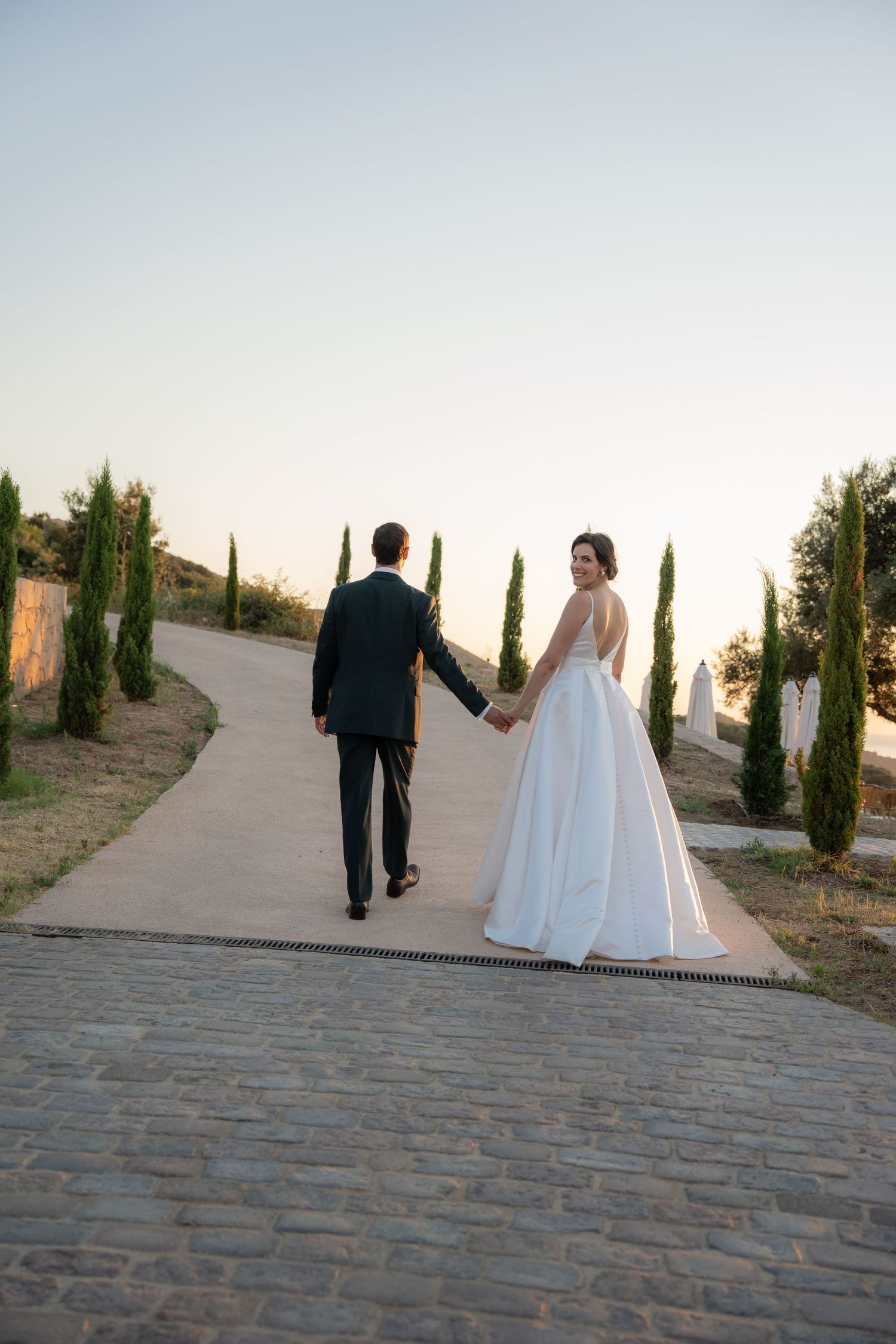 Bride in white gown and groom in dark suit walk hand-in-hand down a path lined with cypress trees at sunset.