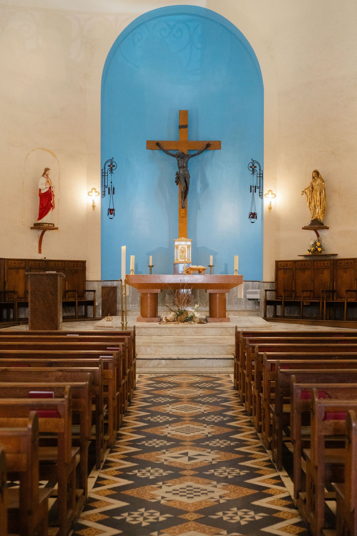 Interior of a Catholic church with blue arched altar, wooden pews, and decorative tiled floor leading to a crucifix.