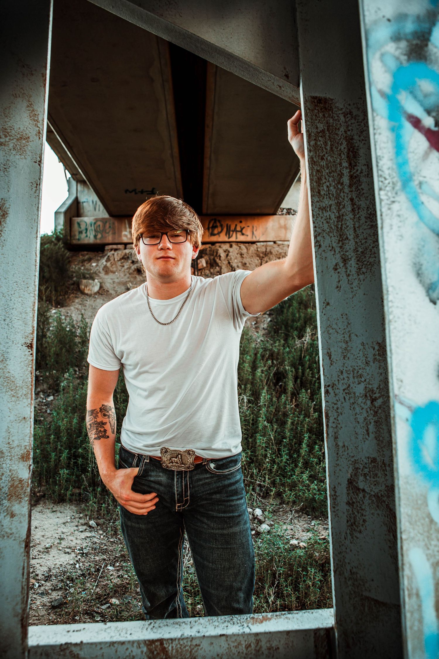 A person in a white t-shirt and jeans poses in an urban doorway with graffiti and stone walls in the background.