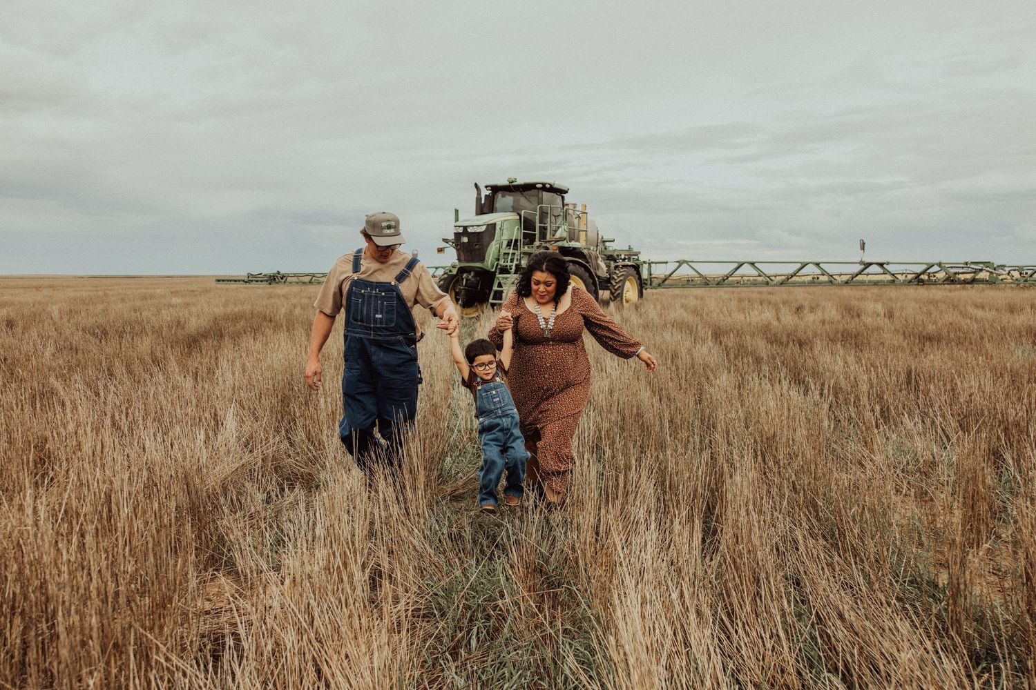 A family walks through a golden wheat field with an agricultural sprayer in the background on an overcast day.