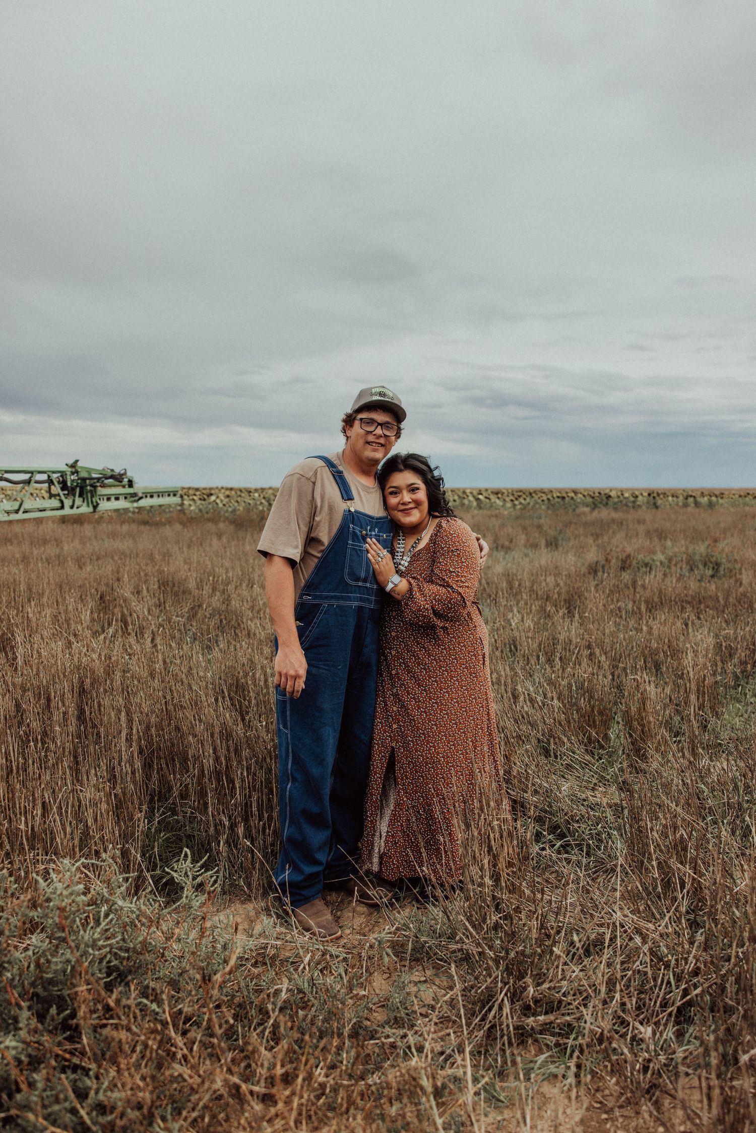 A couple stands together in a brown autumn field under an overcast sky.