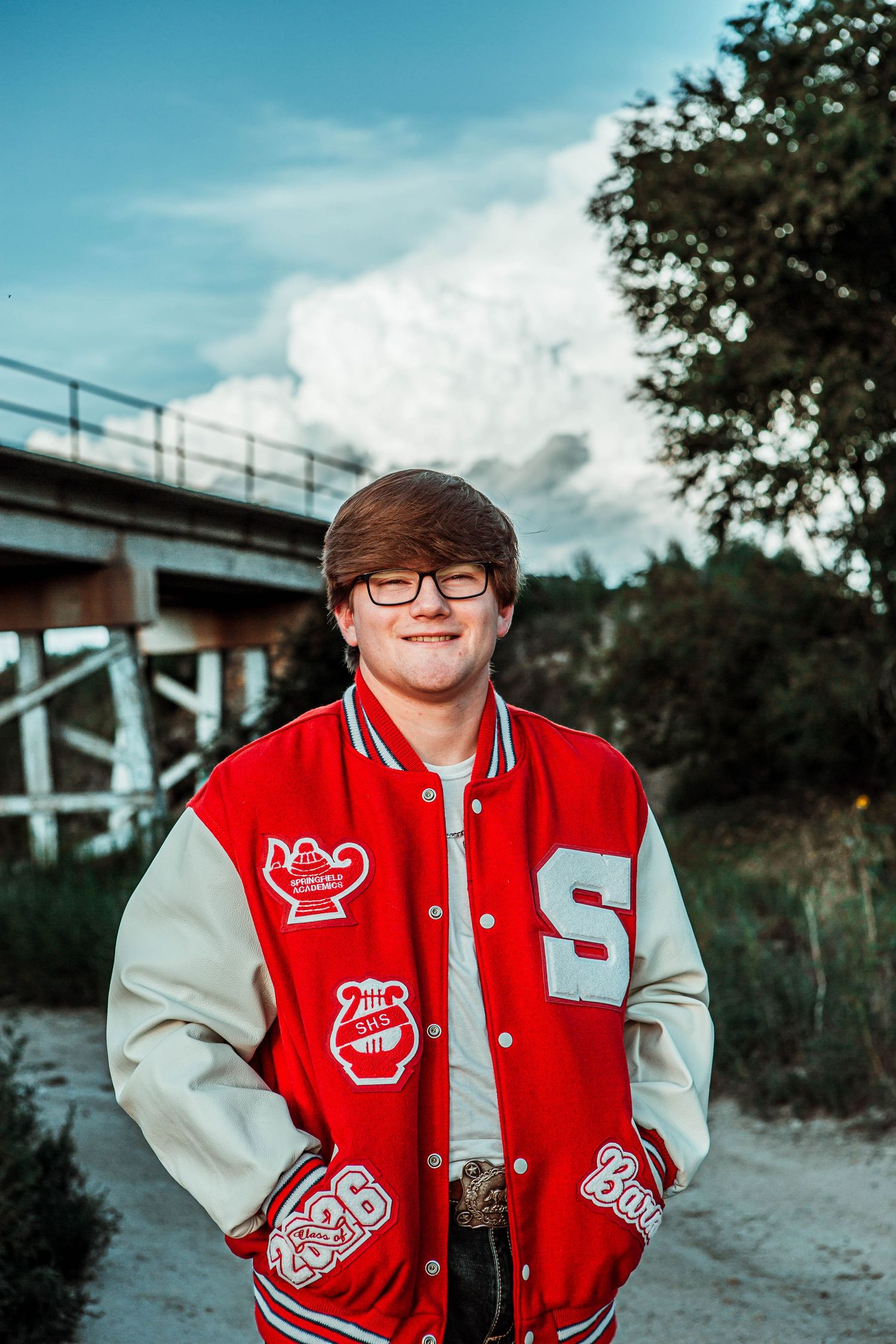A person in a red and gray varsity letterman jacket poses outdoors on a sunny day with trees and buildings in the background.