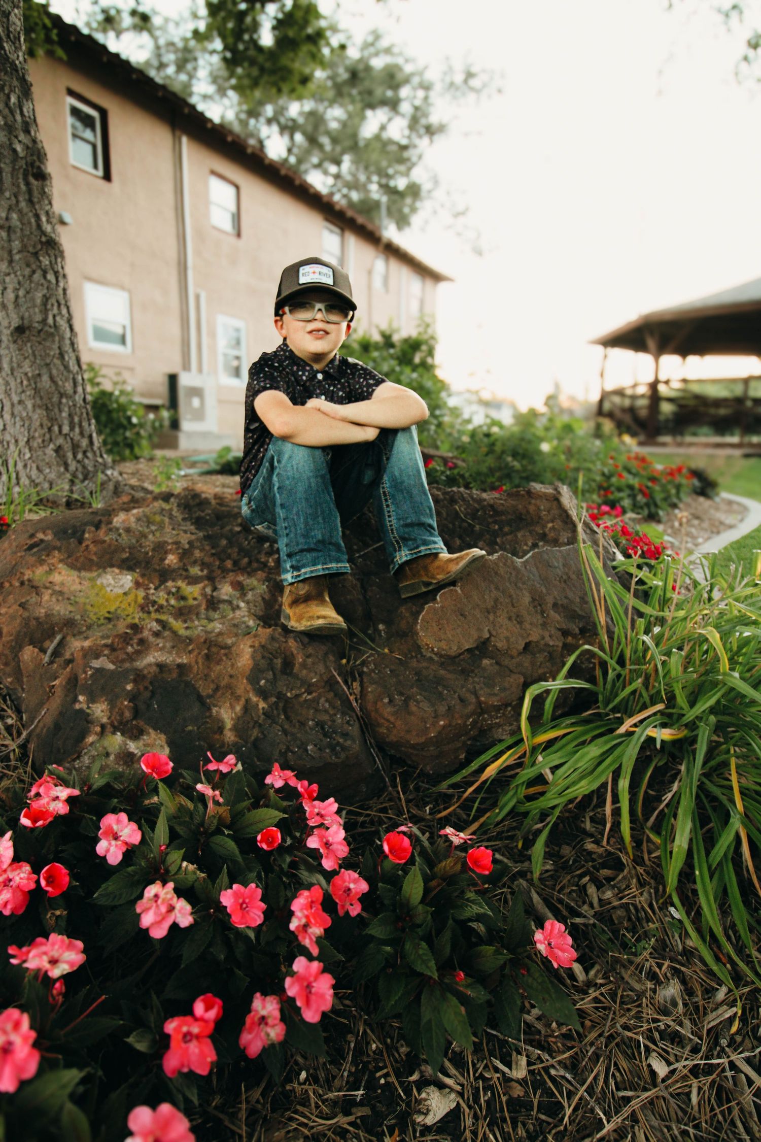 Young person in a baseball cap perches on a rock with pink flowers blooming in the foreground.