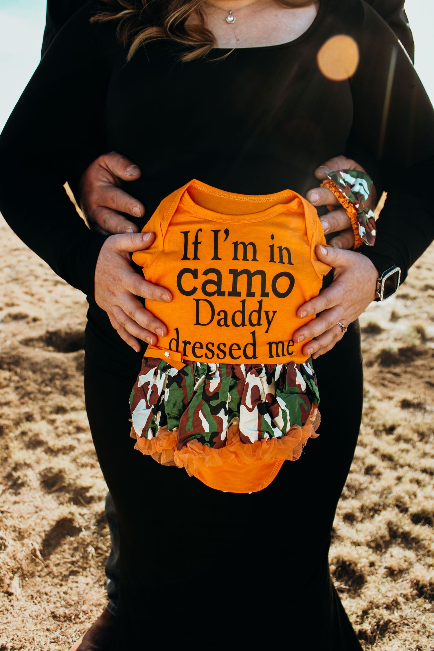 Someone holds up an orange baby onesie with camouflage pattern and text that reads 'If I'm in camo Daddy dressed me'.