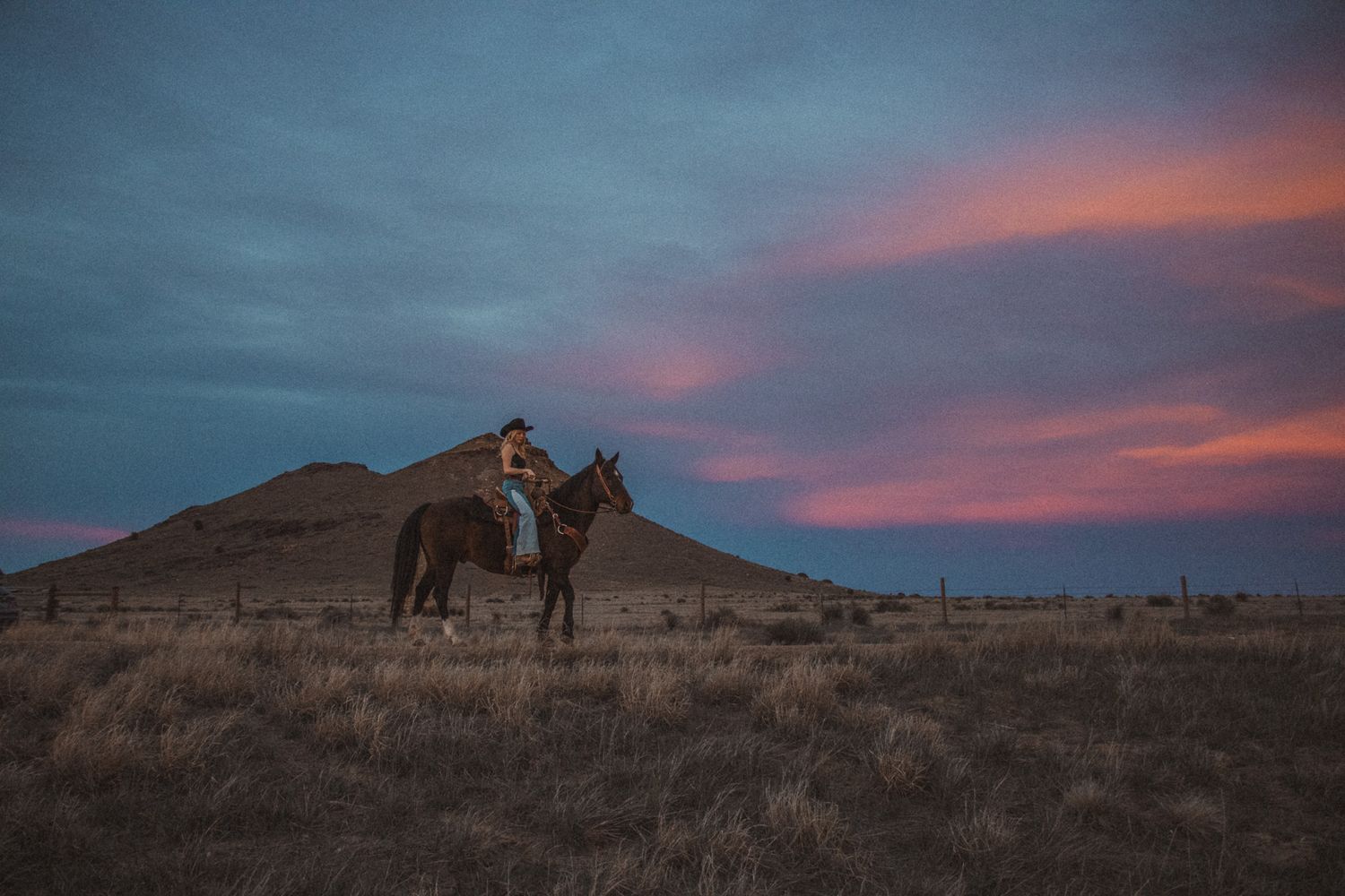A rider on horseback stands silhouetted against a dramatic sunset sky on a hilltop in rugged terrain.