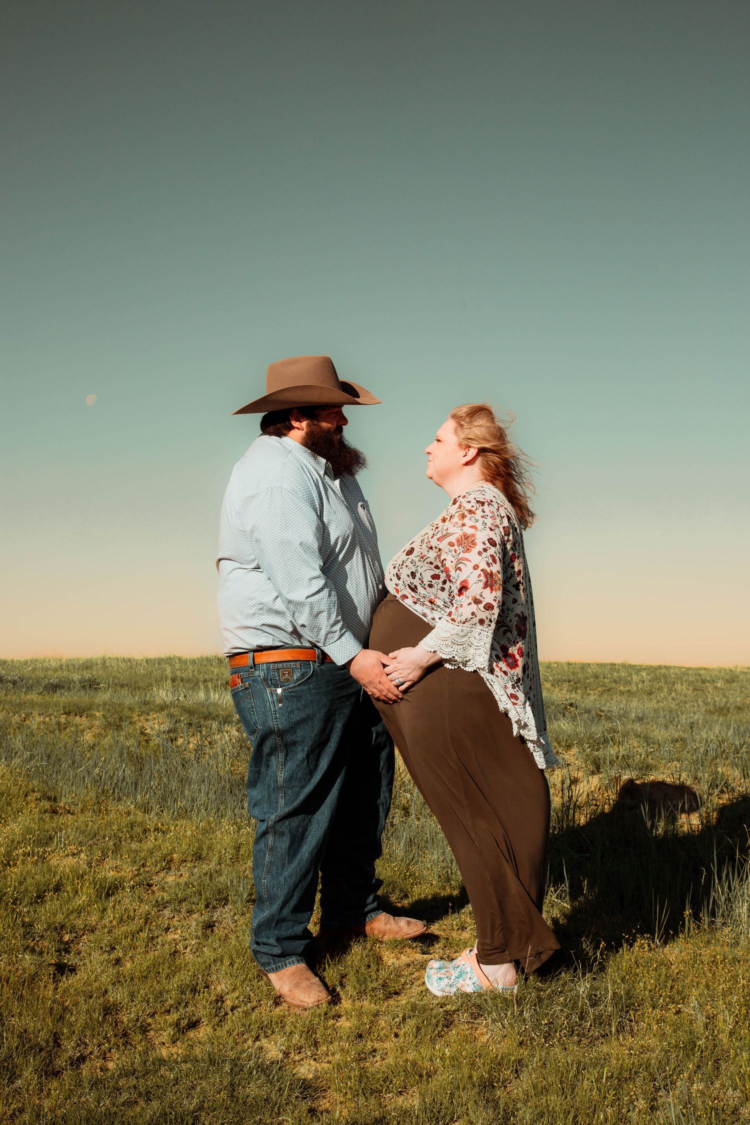 A couple stands together in a grassy field against a pale blue sky during golden hour.