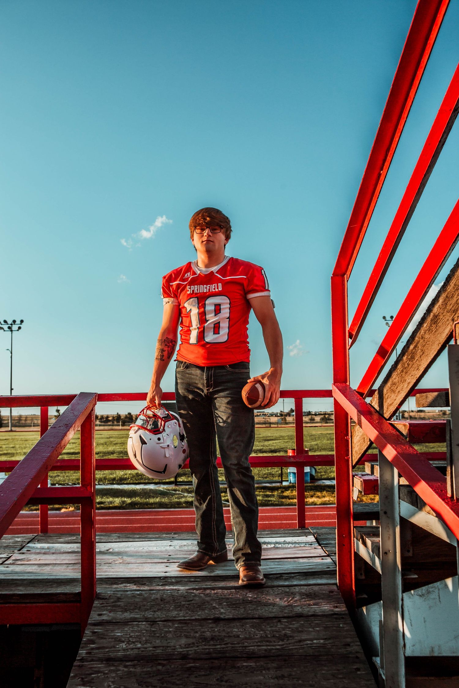 A person sits on red metal bleachers at a sports field during golden hour, creating a striking silhouette against the blue sky.