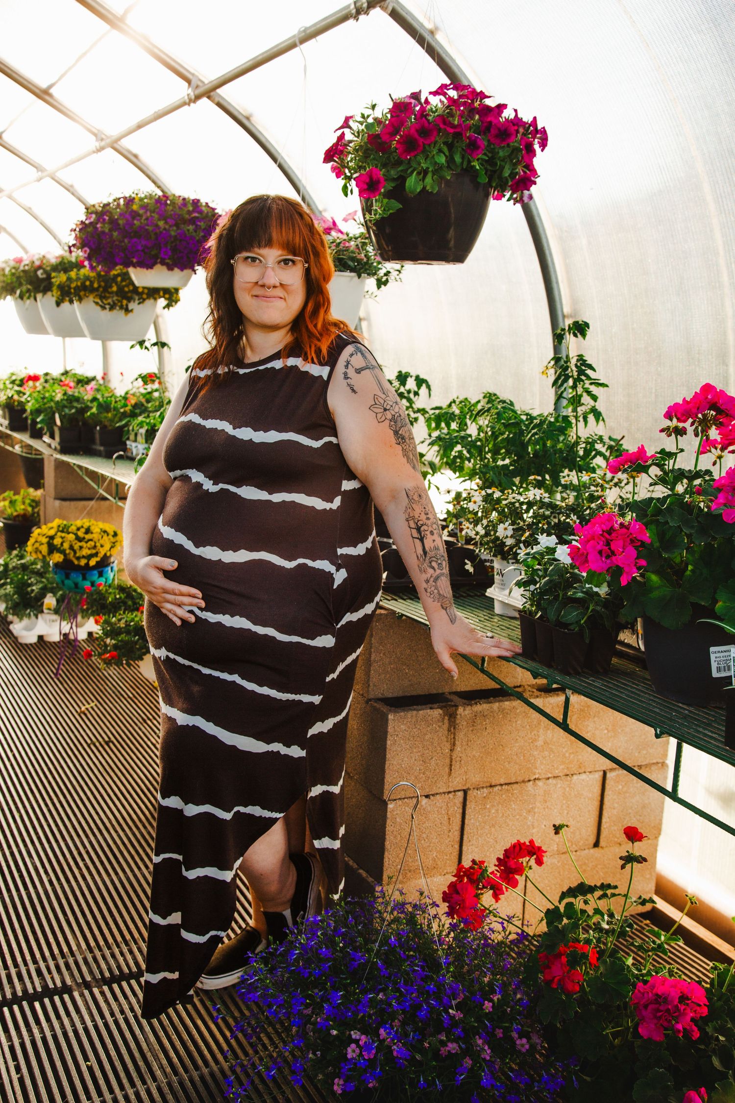 A person in a striped maxi dress stands among colorful flowering plants and hanging baskets in a greenhouse setting.