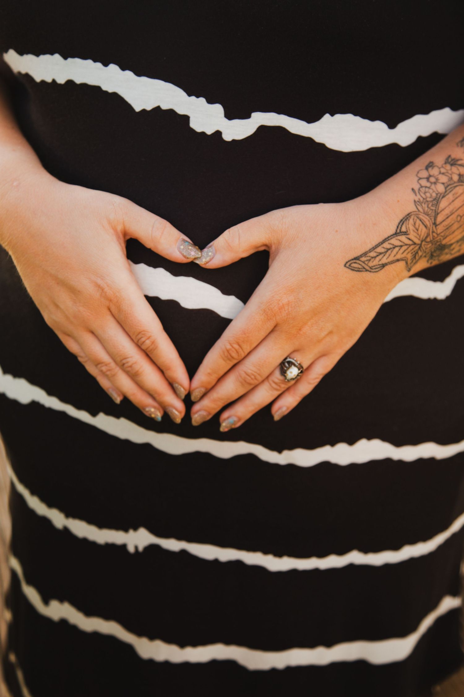 Hands forming a heart shape against a black and white striped background, showing tattoos and a gemstone ring.